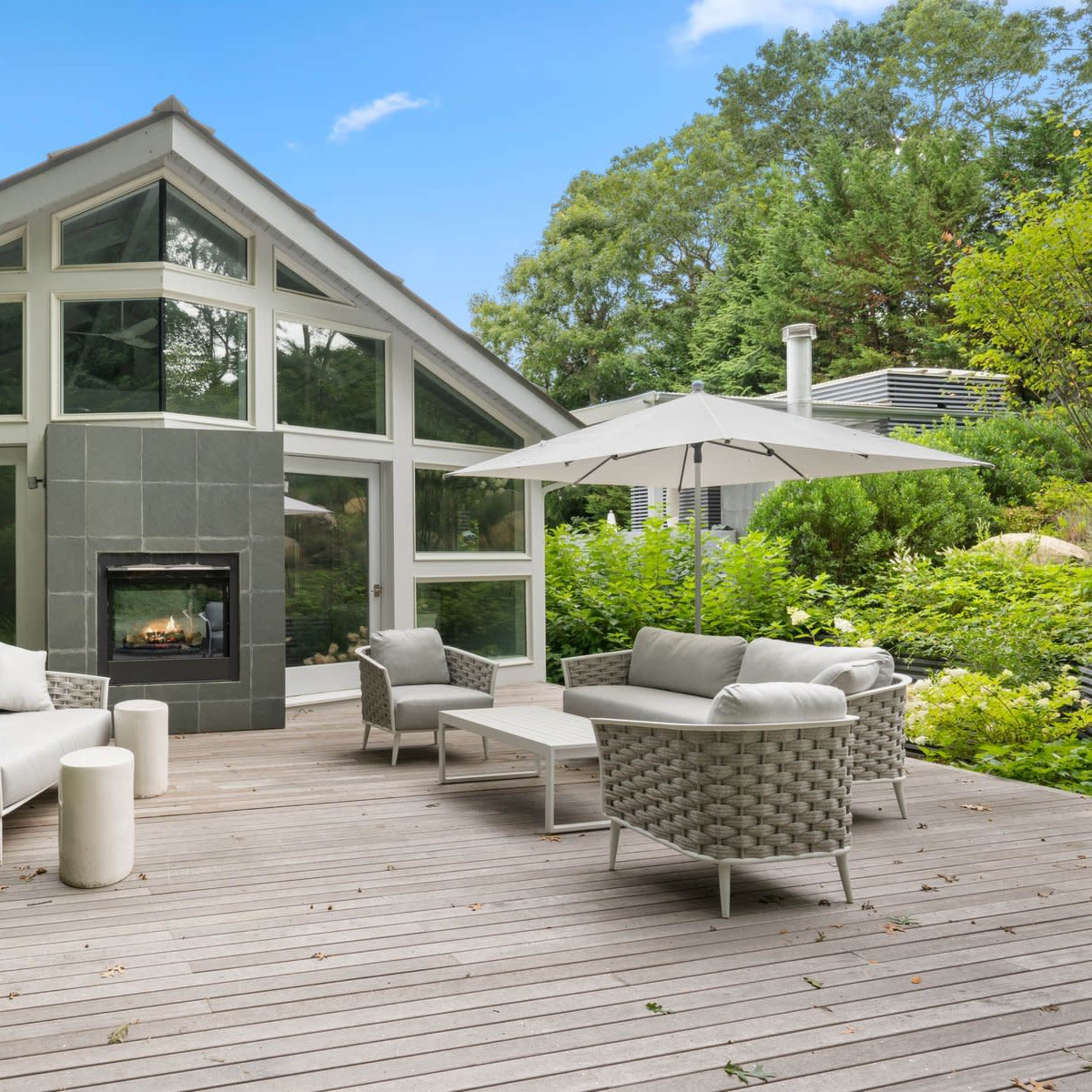 A modern deck features comfortable seating arrangements and a patio umbrella in front of a large glass-paneled house surrounded by lush greenery.
