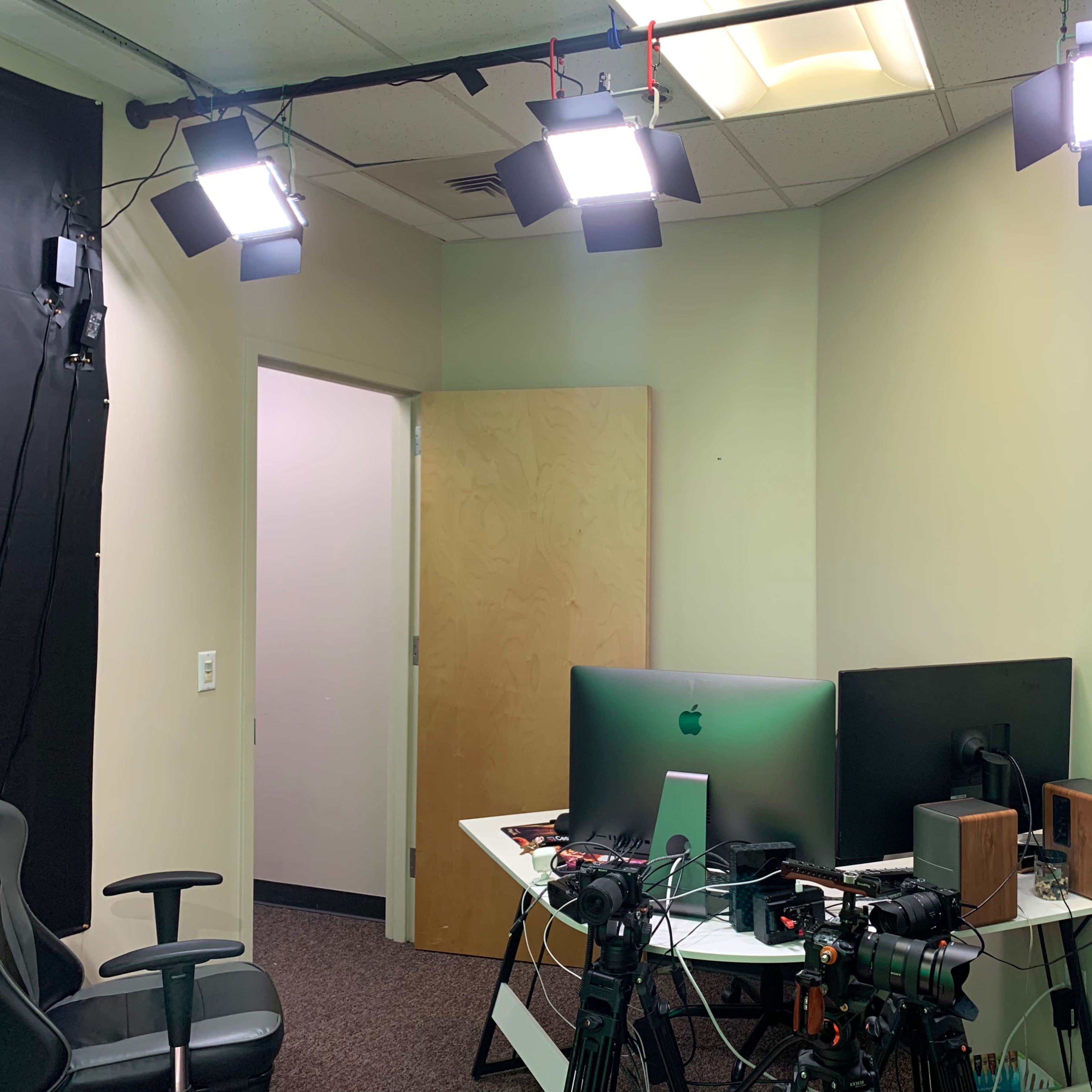 A dimly lit room with three ceiling lights illuminating a desk containing two computer monitors and cameras, while a door is visible in the background.