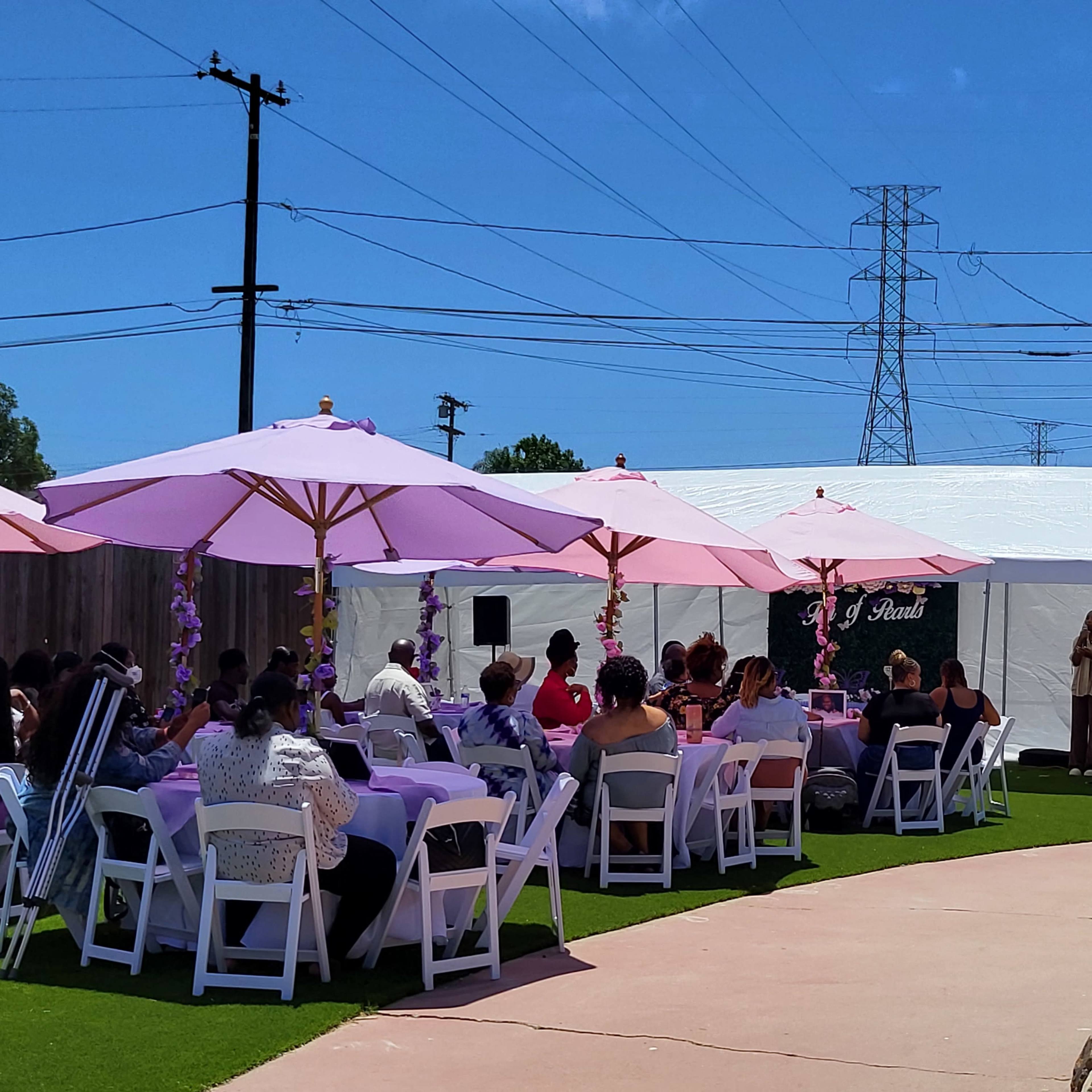 A large outdoor gathering takes place under pink umbrellas, with seated guests around tables and a musician performing near a white tent.