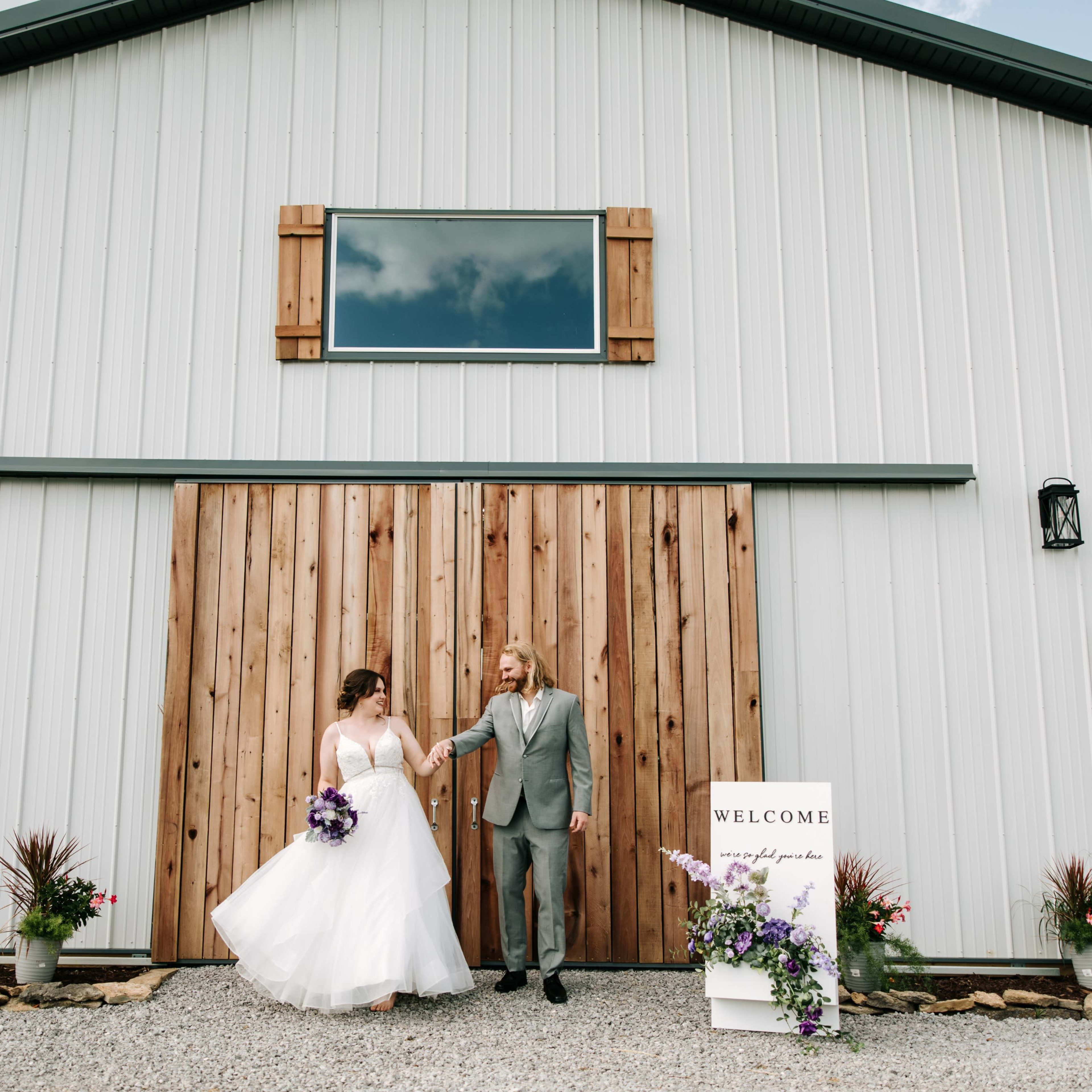 A bride and groom walk hand in hand in front of a modern barn venue adorned with wooden accents and flower arrangements.