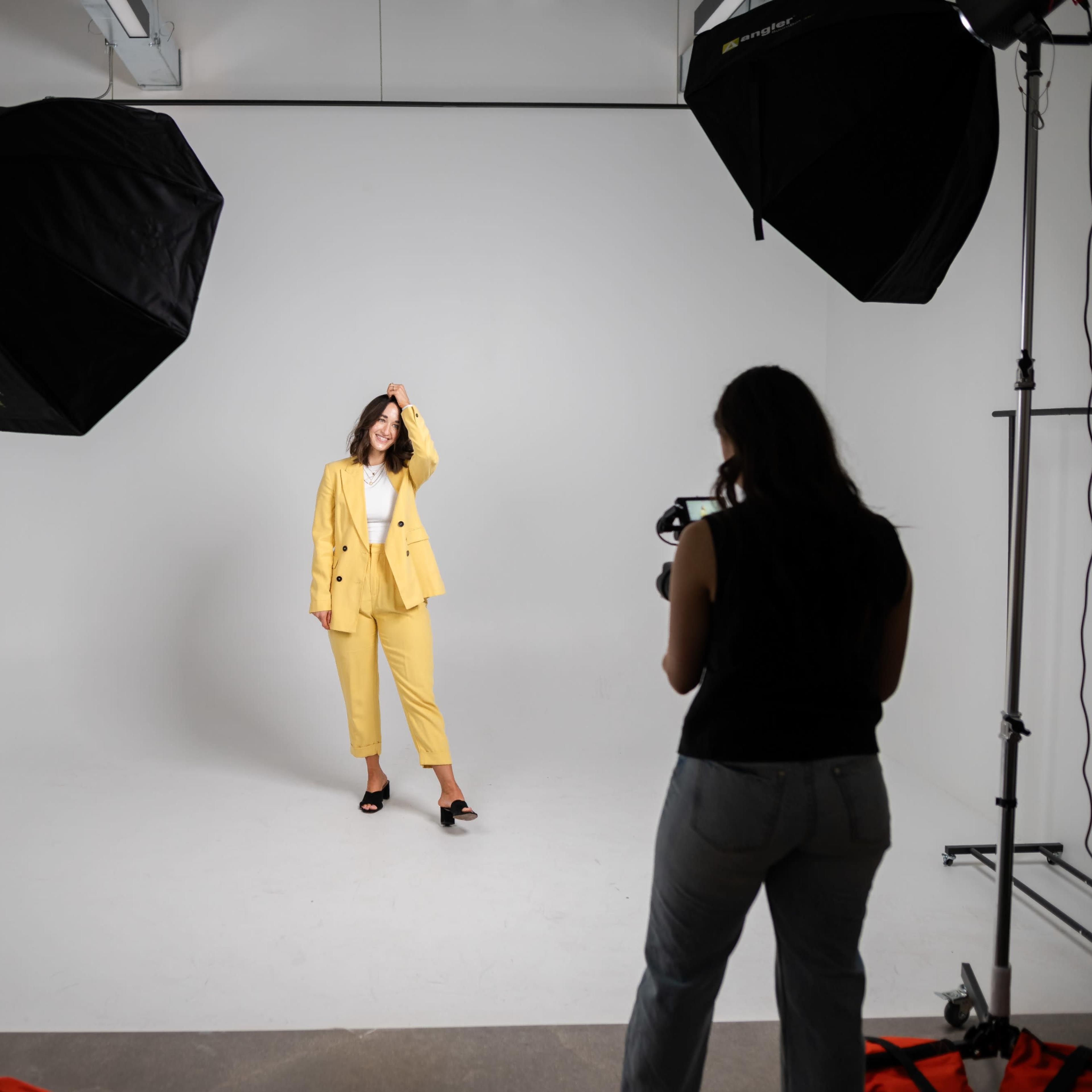 A woman in a yellow suit poses in a studio while another woman takes her picture with a camera.
