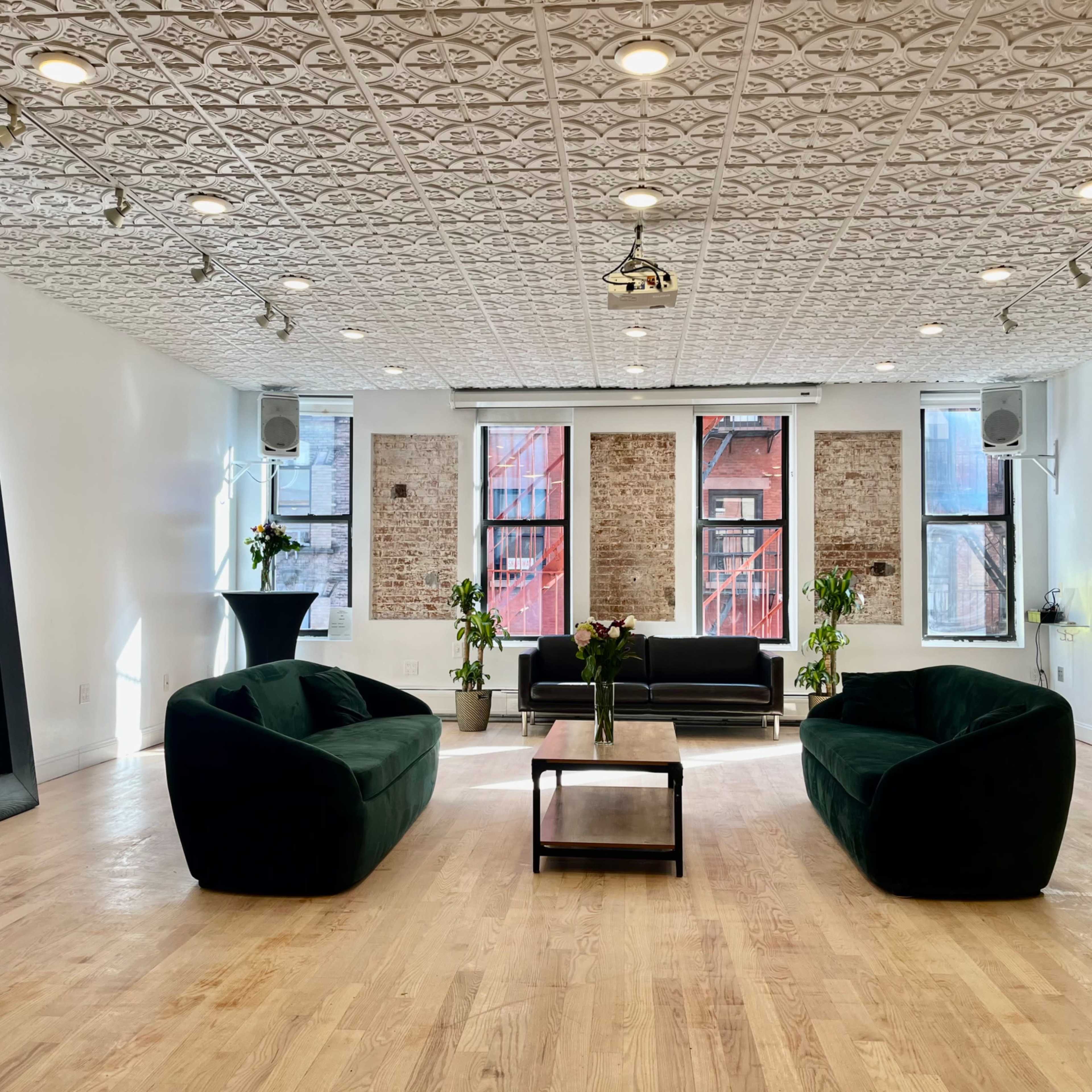 The room features two green sofas, a black couch, a coffee table, and a mirror, all set against a backdrop of exposed brick walls and bright windows.