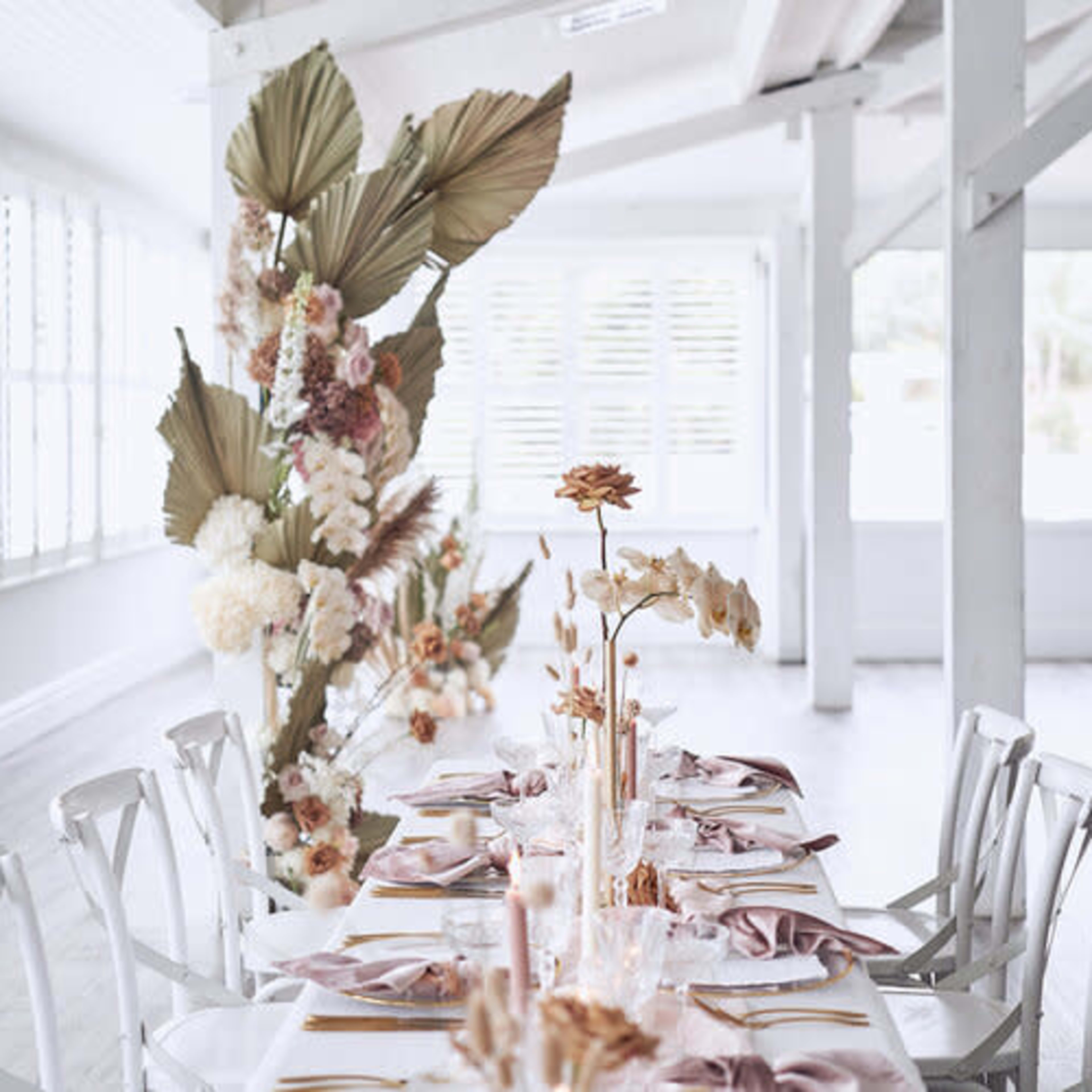 A long dining table is elegantly set with decorative flowers and napkins in a brightly lit, minimalist room.
