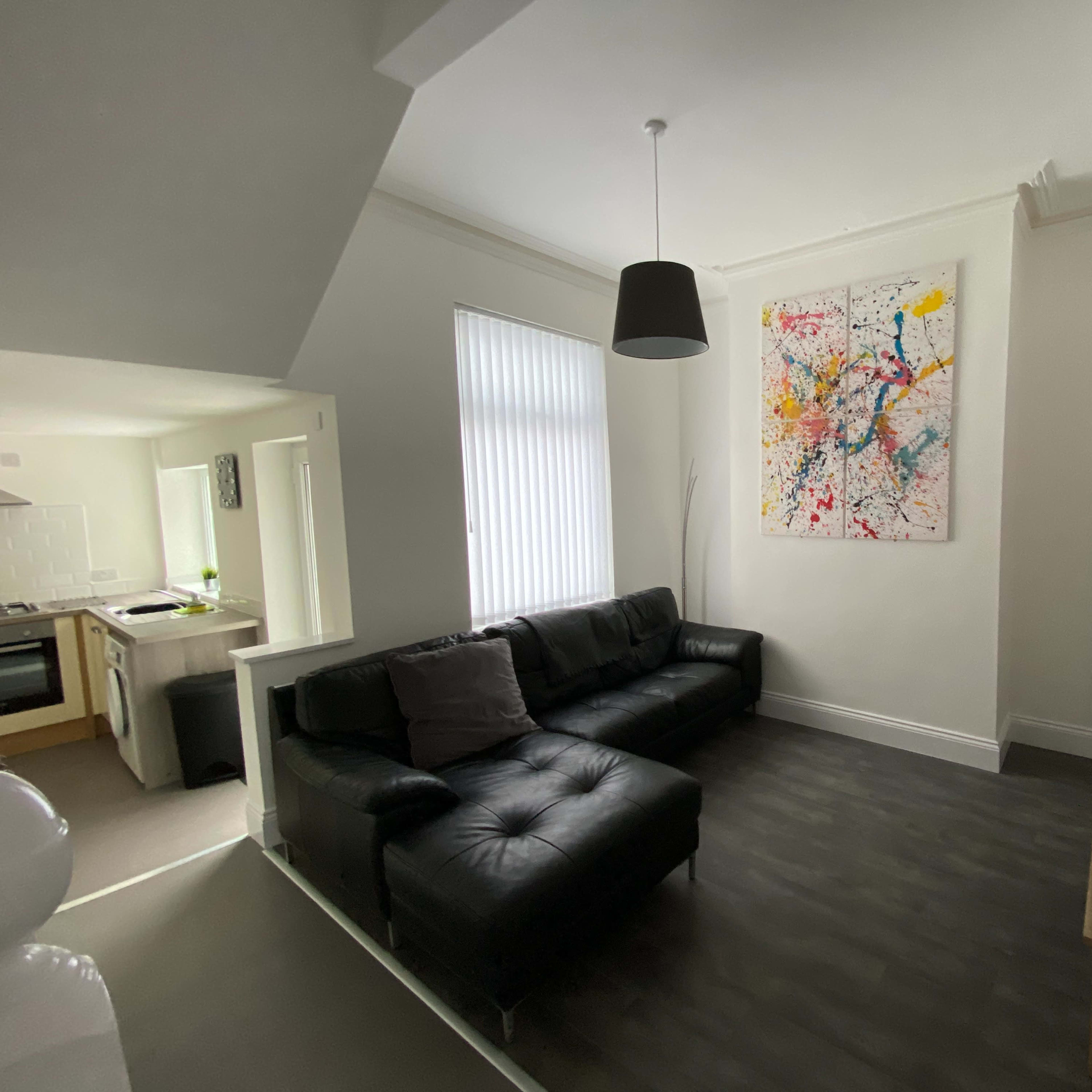 A modern living room features a black leather sofa, a wall-mounted abstract painting, and a small wooden television stand.