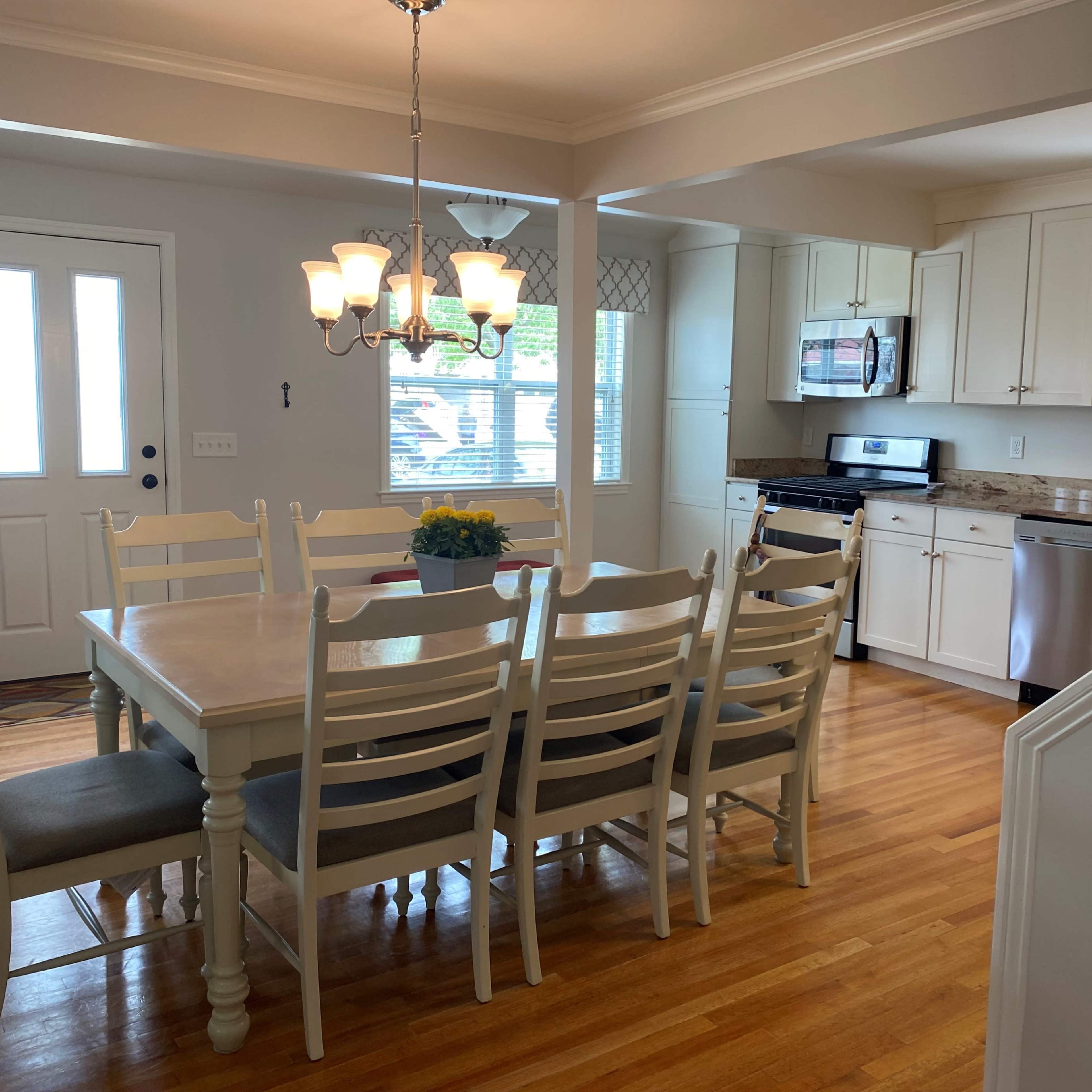 A well-lit dining area featuring a large white wooden table surrounded by chairs, with a view of a modern kitchen and an entrance door in the background.