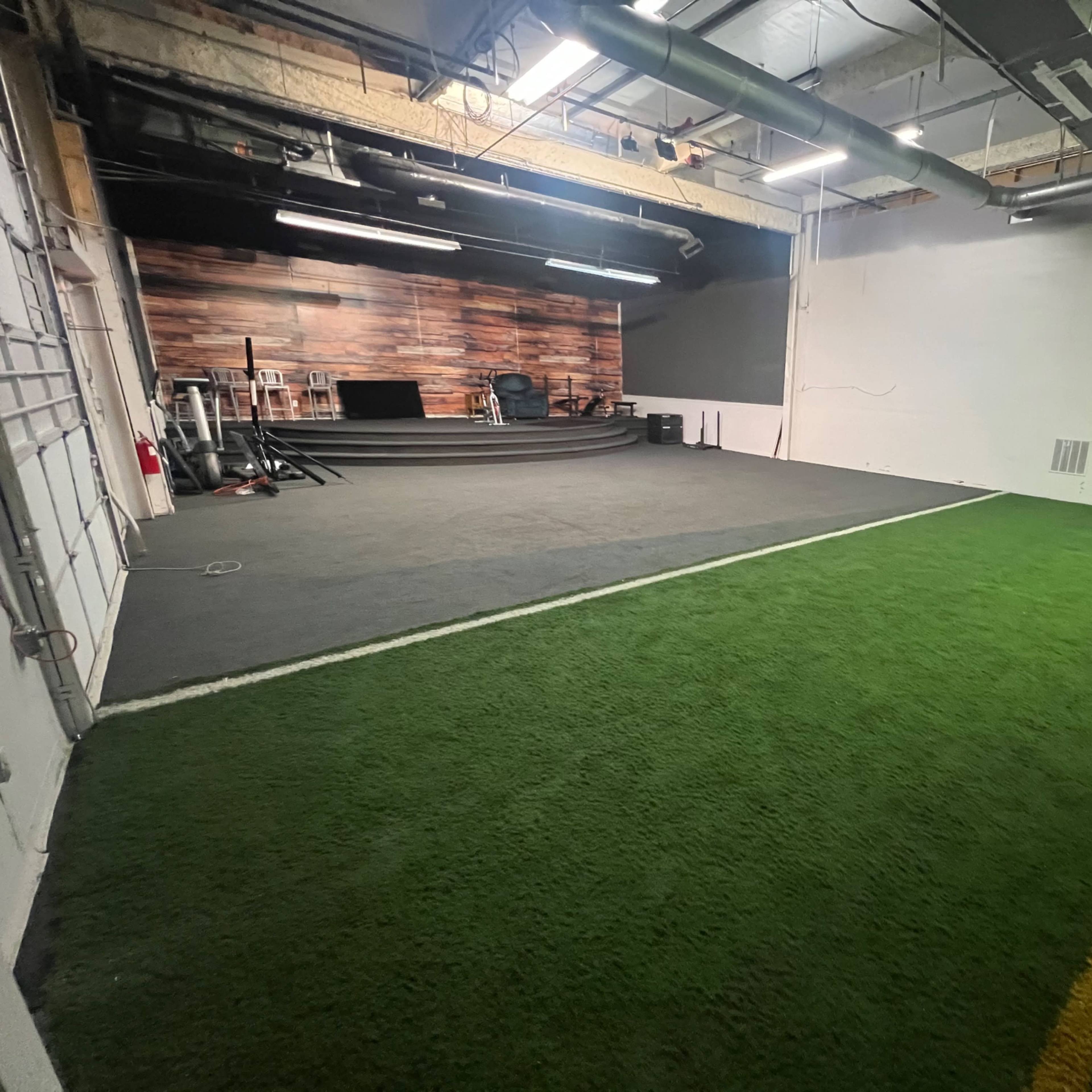 A spacious indoor training facility with a section of artificial turf and a wooden-paneled wall in the background.