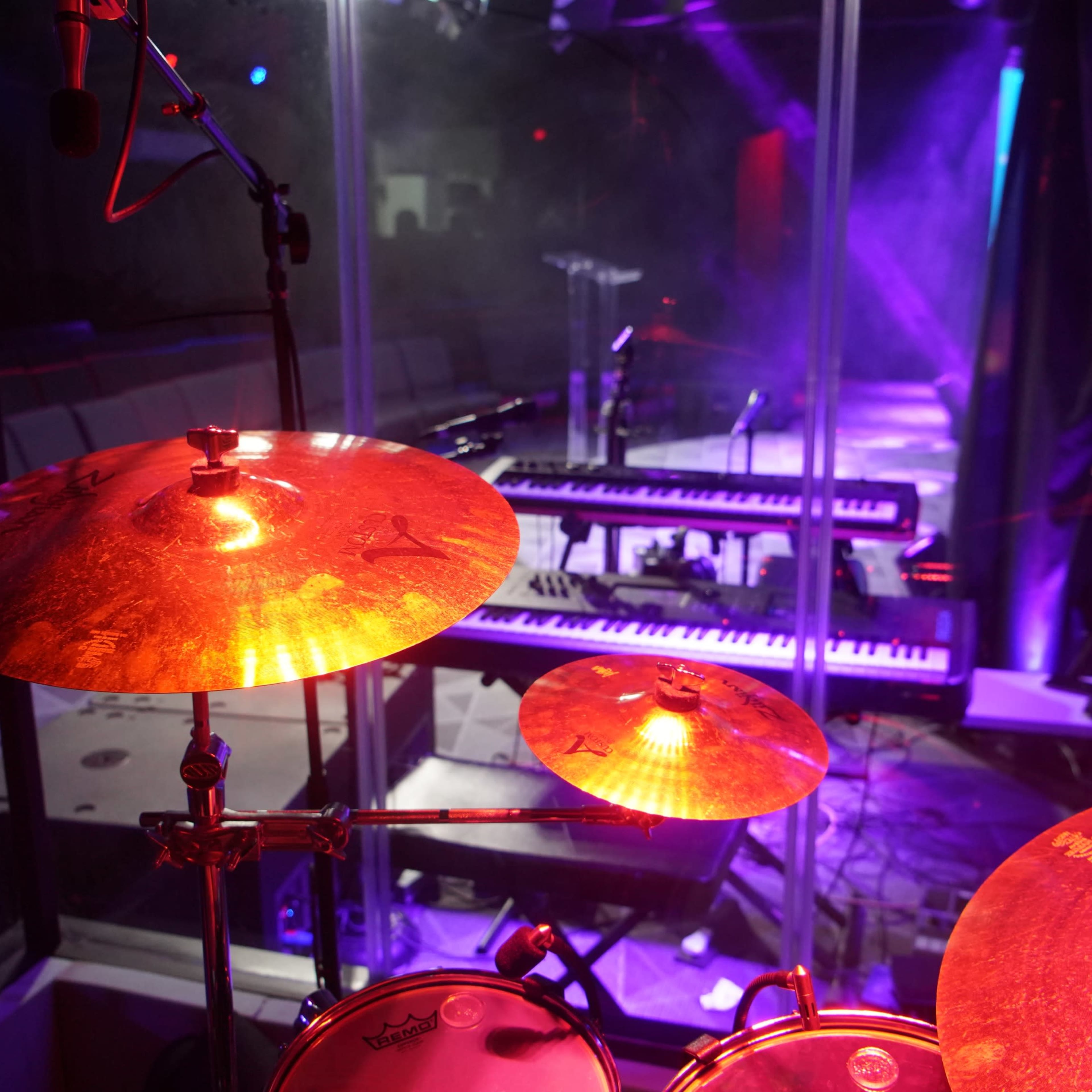 A view from behind a drum set, highlighting illuminated cymbals and a keyboard setup in a music performance space surrounded by glass.