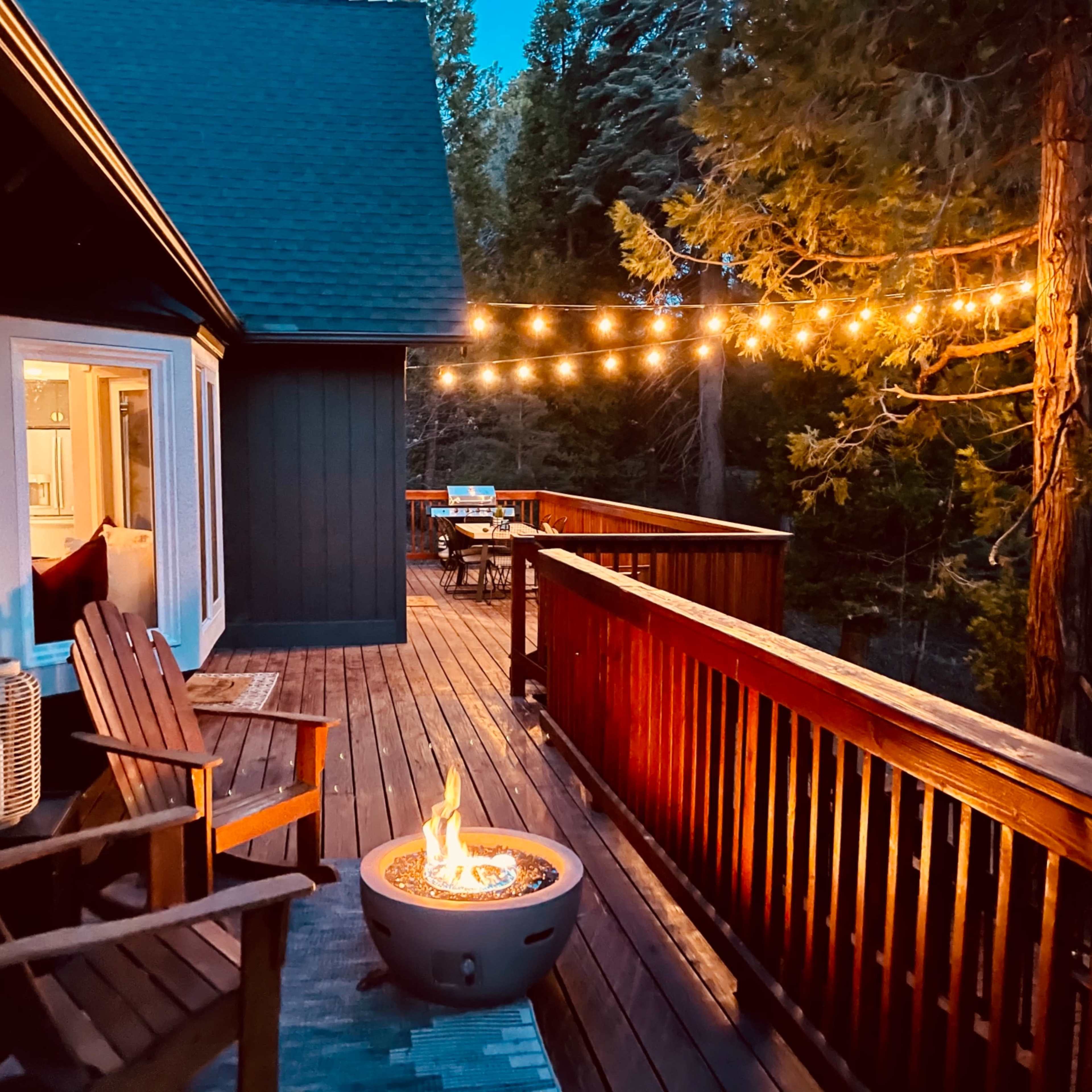 A wooden deck features a fire pit surrounded by chairs, string lights overhead, and a view of trees in the evening.