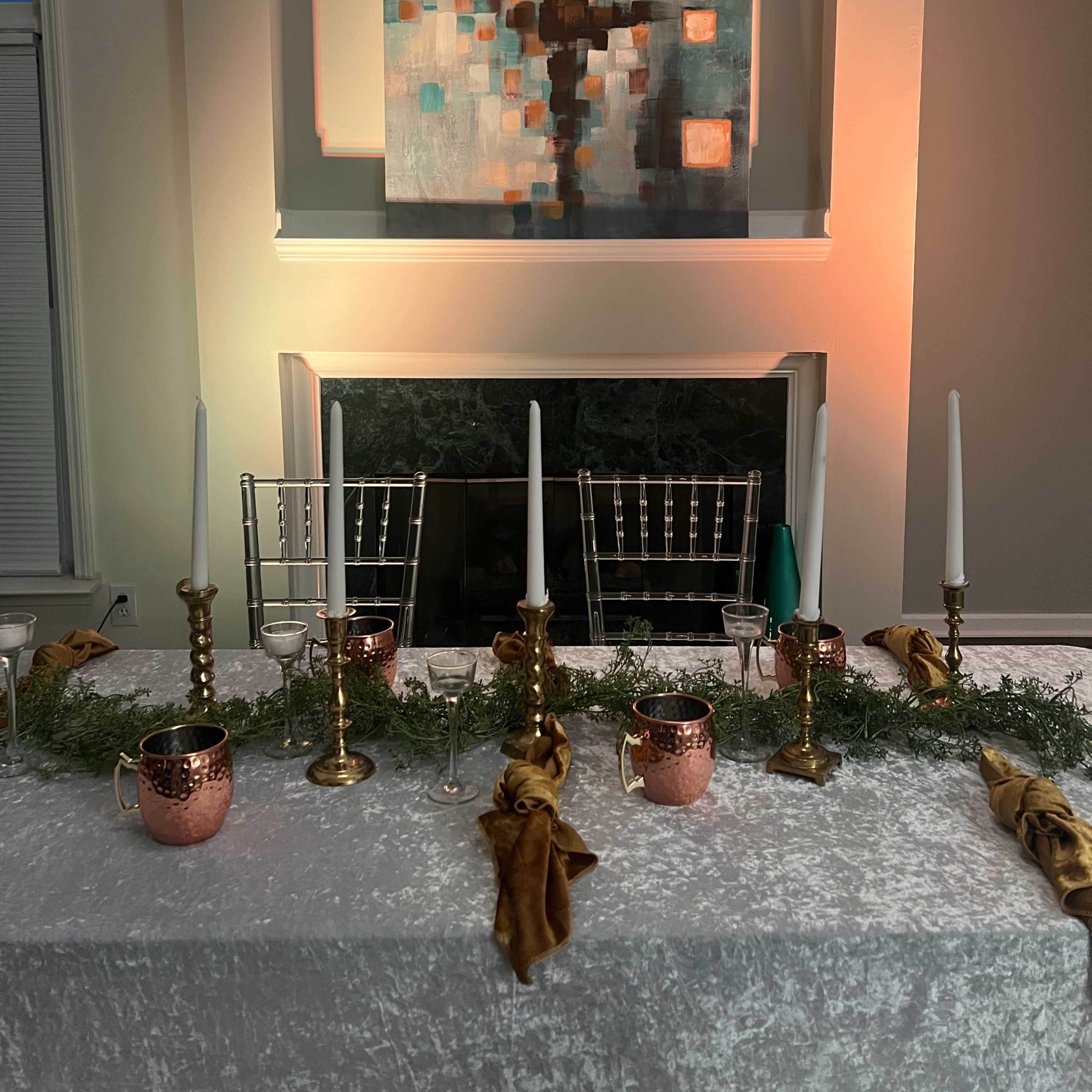 A dining table is set with gold and copper decor, white candles, and greenery in front of a modern abstract painting and a lit fireplace.