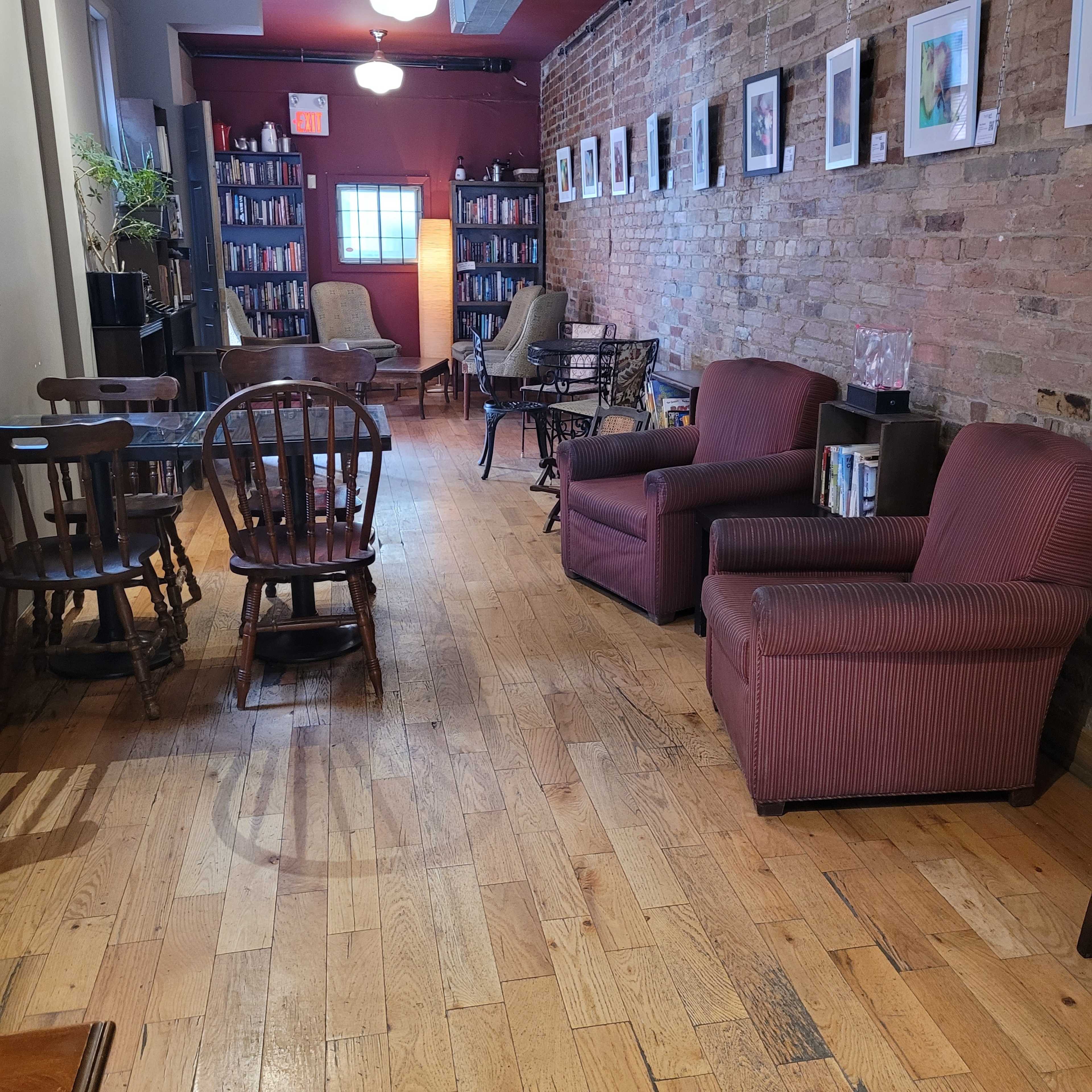 A cozy interior of a small library or reading area with wooden floors, bookshelves lined with books, and seating arrangements consisting of chairs and small tables.