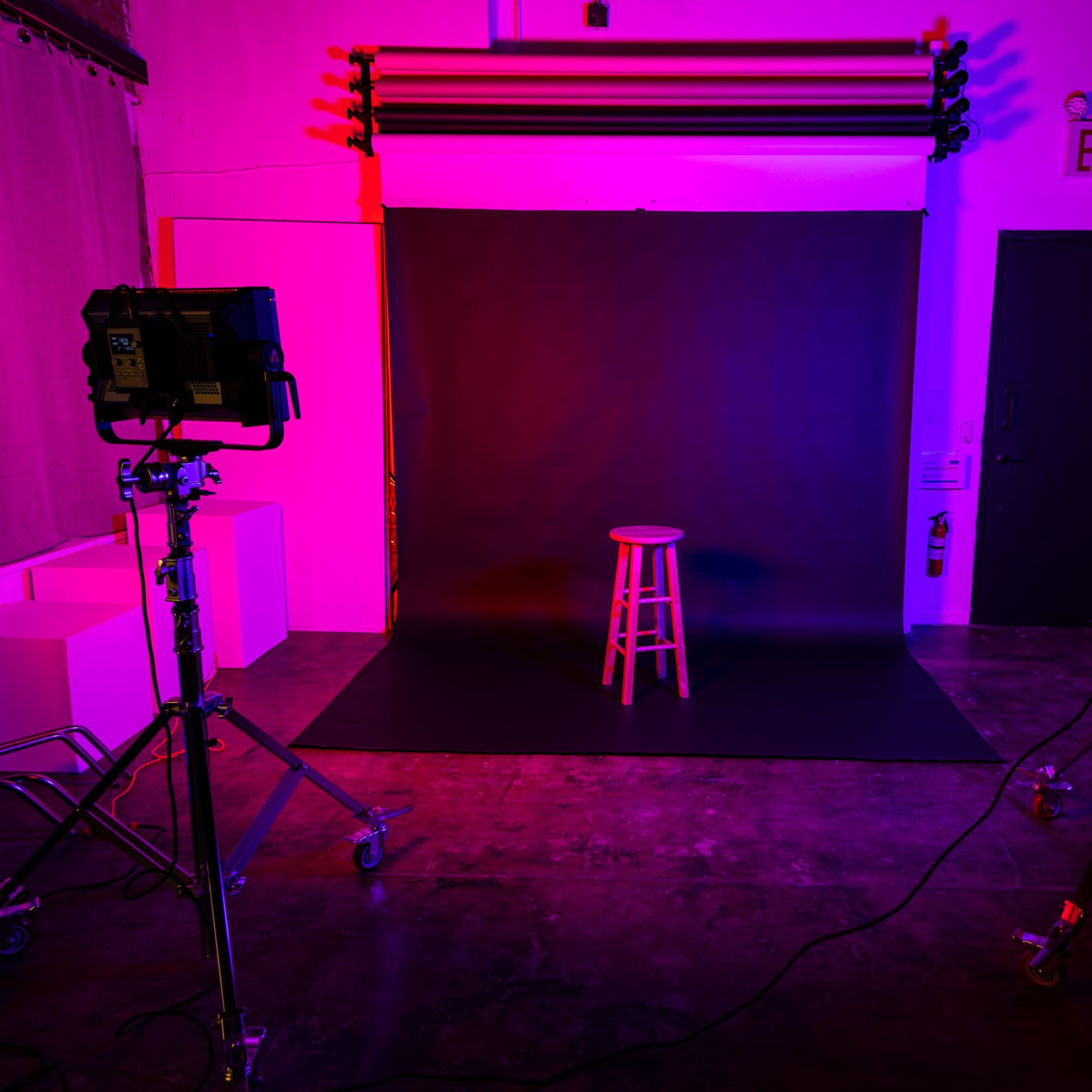 A photography studio features a black backdrop with a wooden stool centered and illuminated by colored lights from two stands on either side.