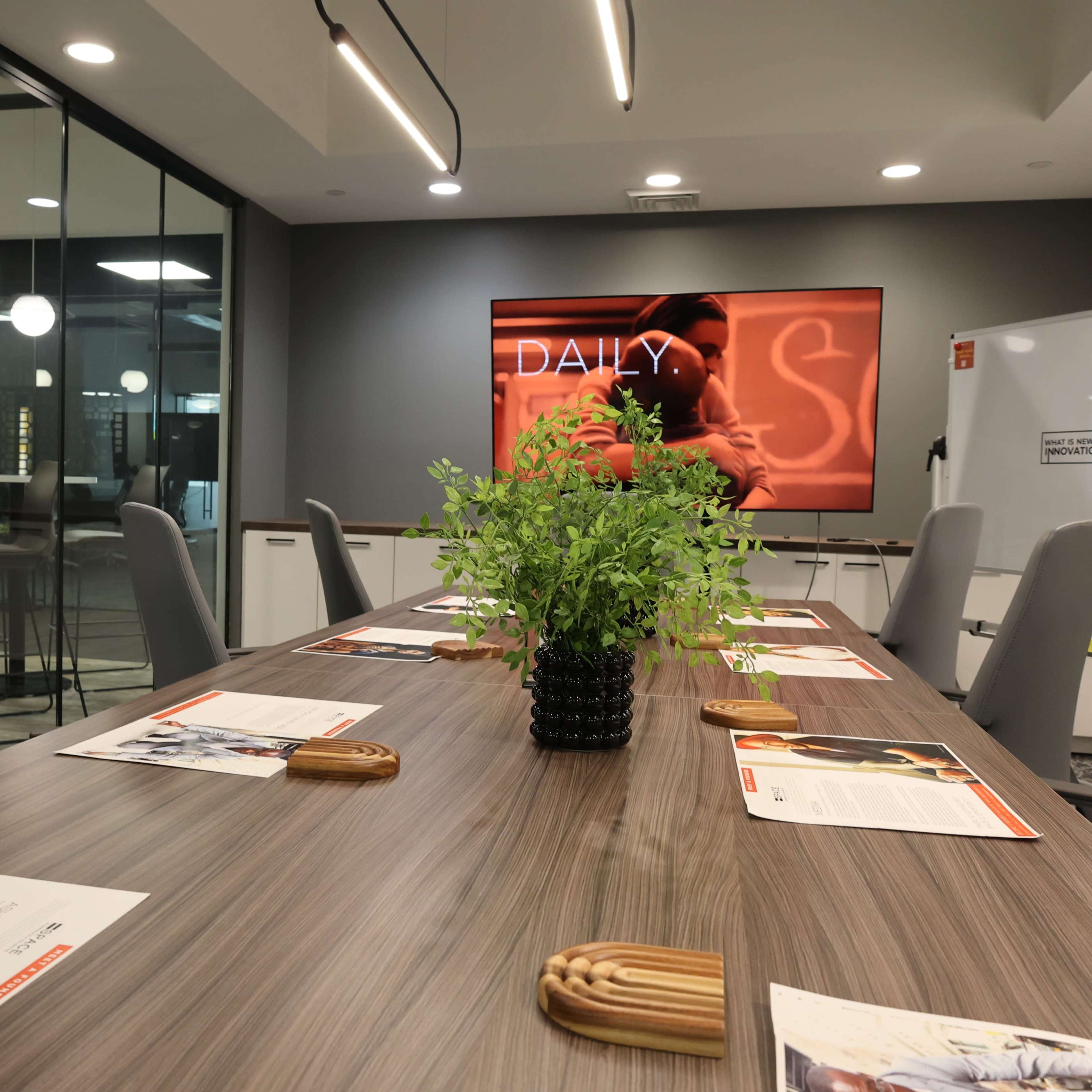 A modern conference room features a long wooden table surrounded by gray chairs, with a large screen displaying the word "DAILY" and a potted plant as the centerpiece.