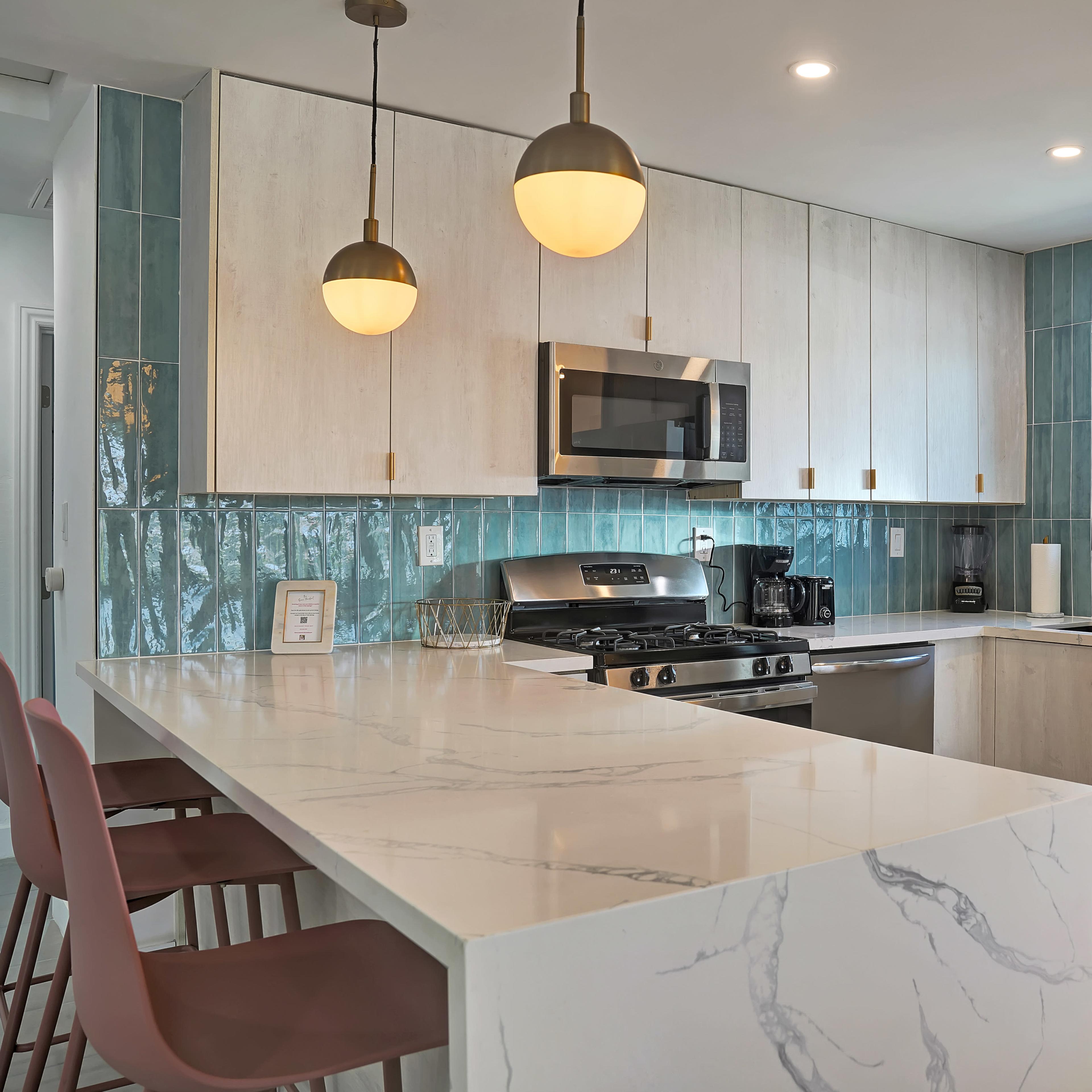 A modern kitchen featuring light-colored cabinetry, a marble countertop with bar stools, stainless steel appliances, and teal tiles on the backsplash.