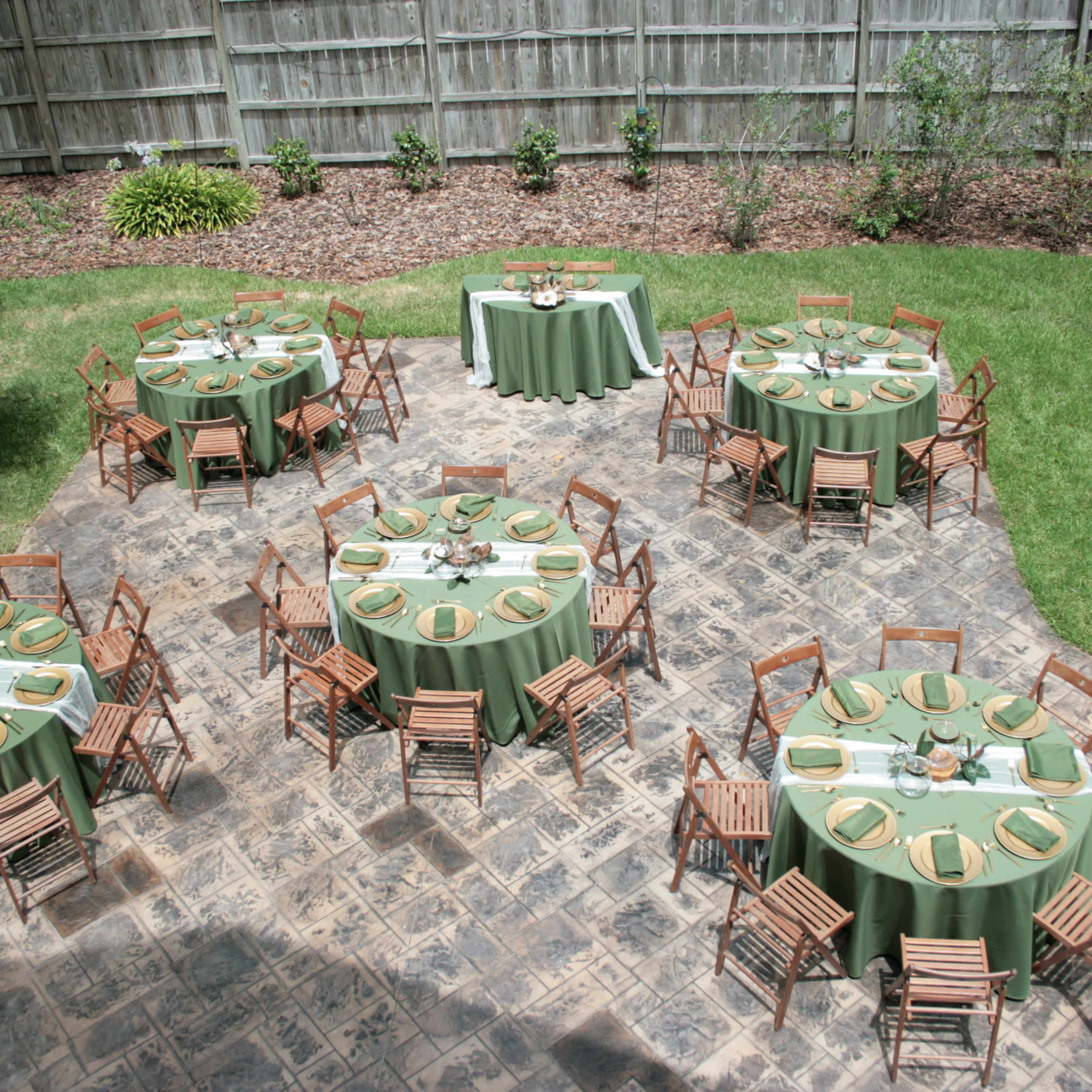 An outdoor event setup with several round tables covered in green linens, each surrounded by wooden chairs, arranged on a stone patio amidst a landscaped yard.