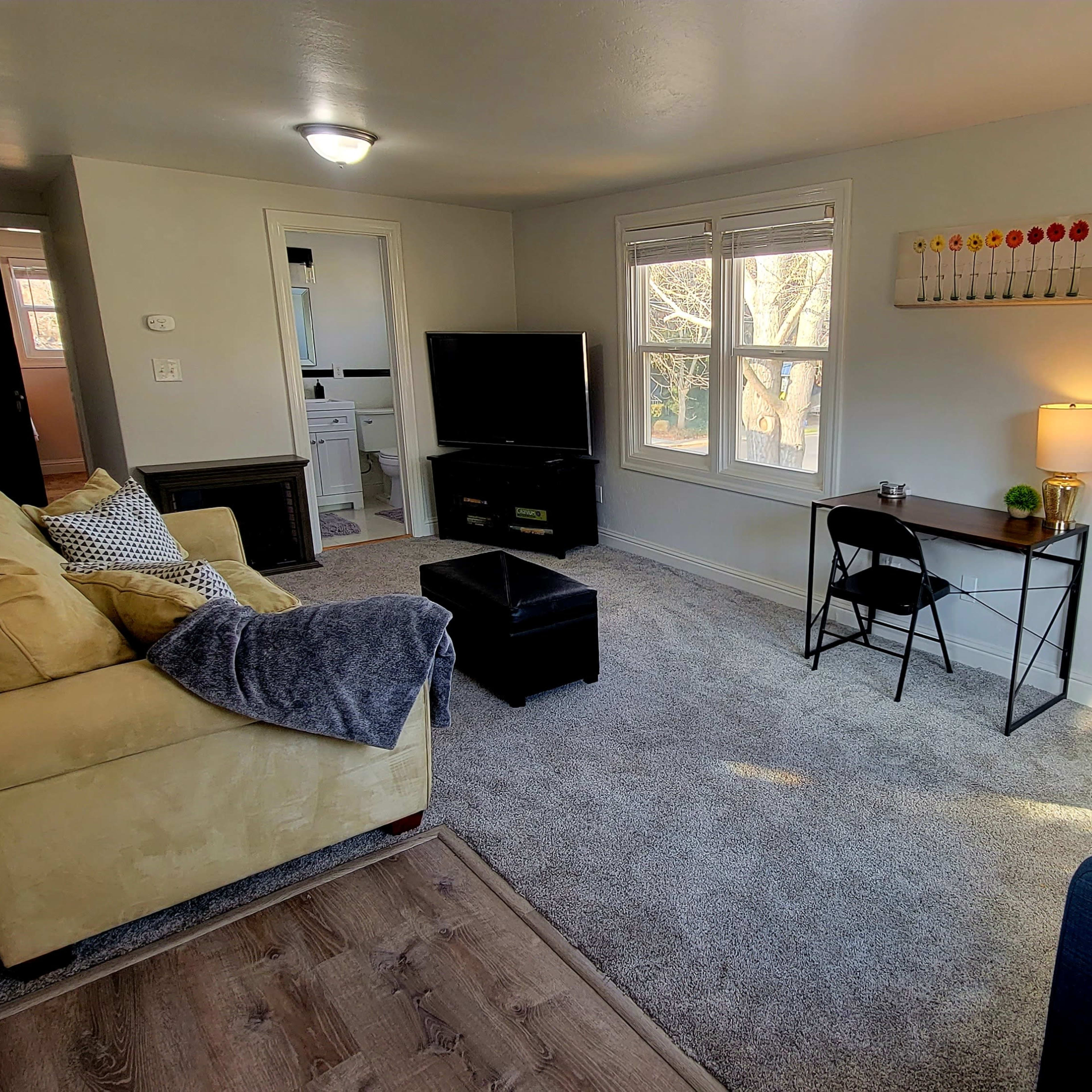 The living room features a yellow sofa, a blue chair, a coffee table, a small desk with a lamp, and a television against a light-colored wall.