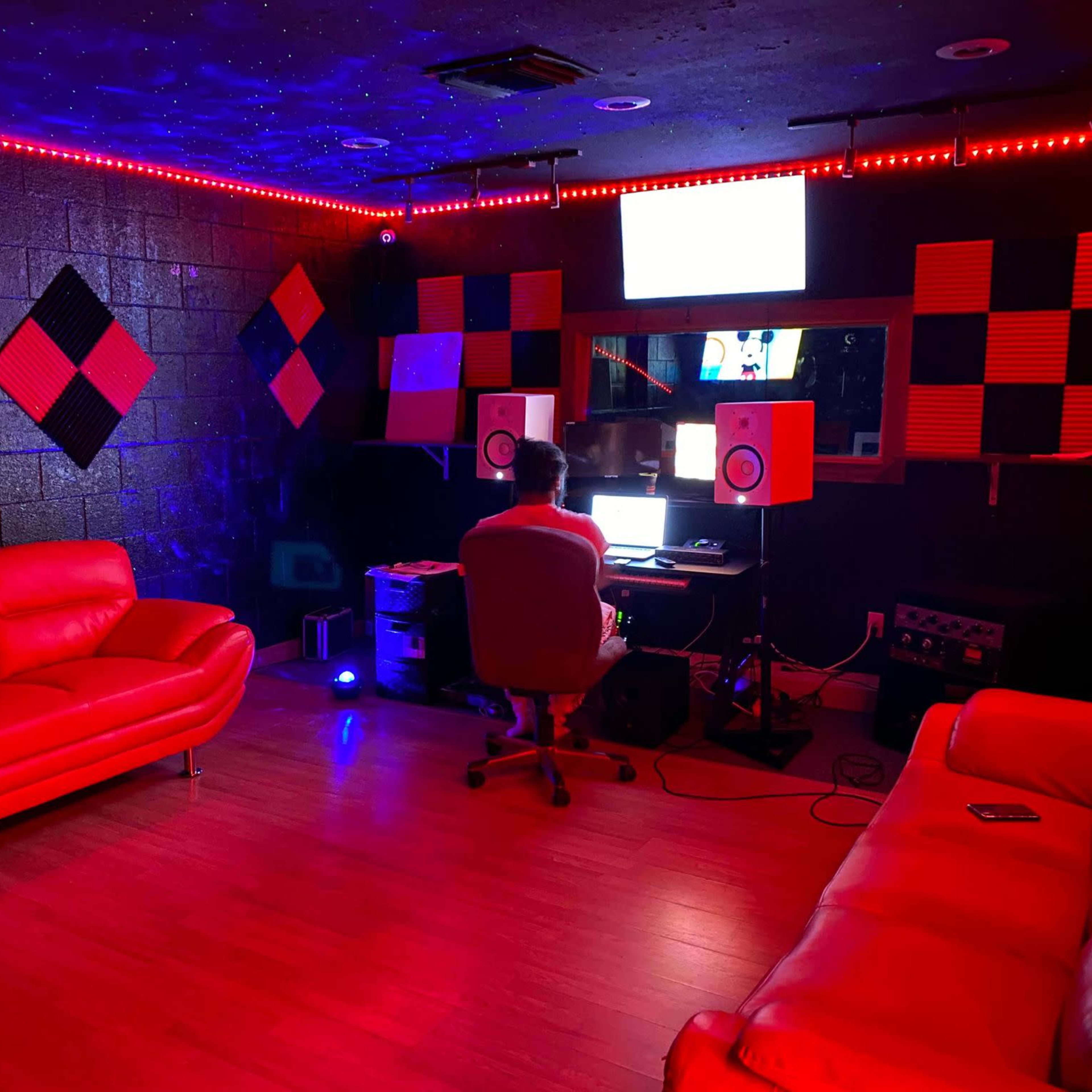 A person sits at a computer in a music studio furnished with red couches, colorful sound panels, and ambient lighting.