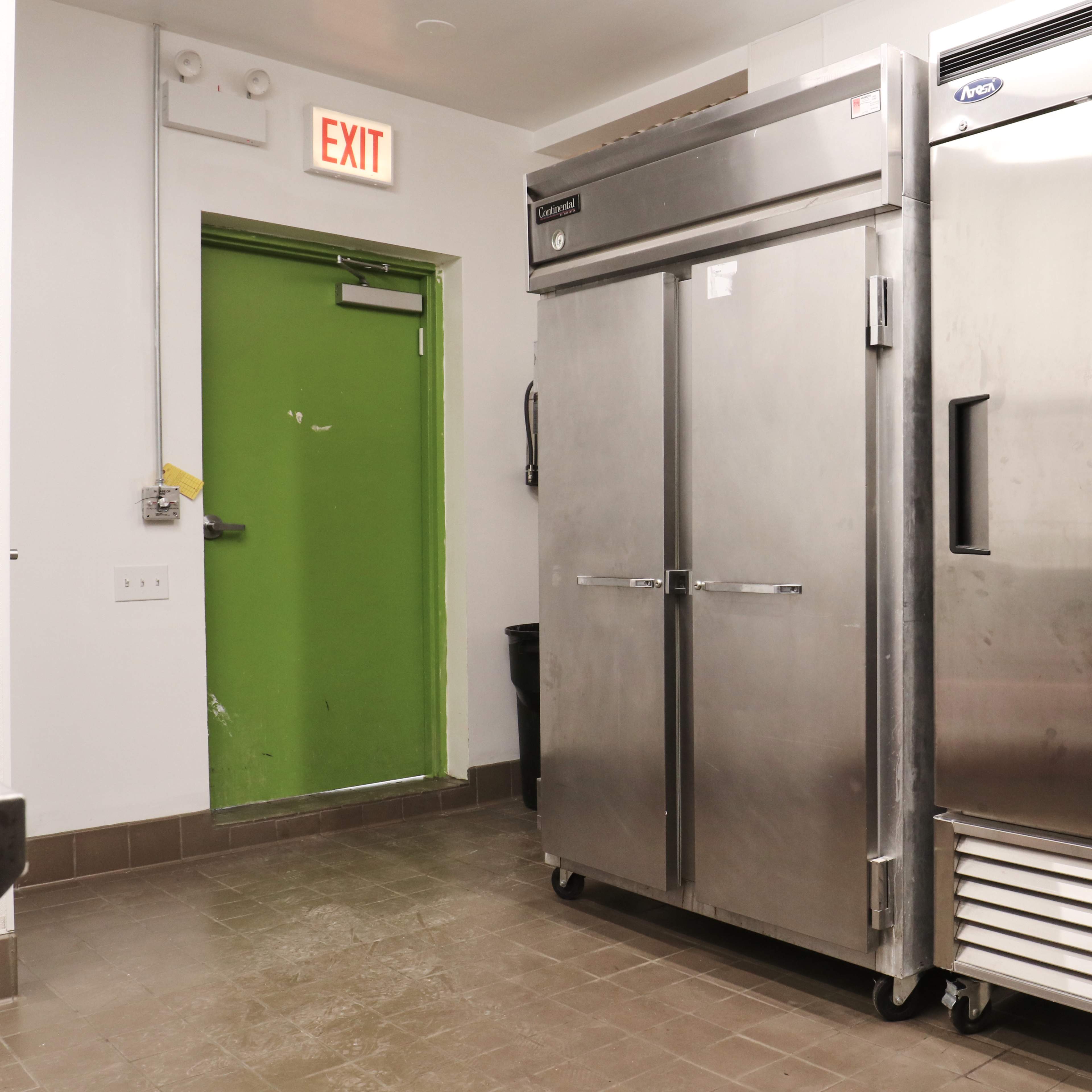 Two stainless steel refrigerators next to a green door in a commercial kitchen.