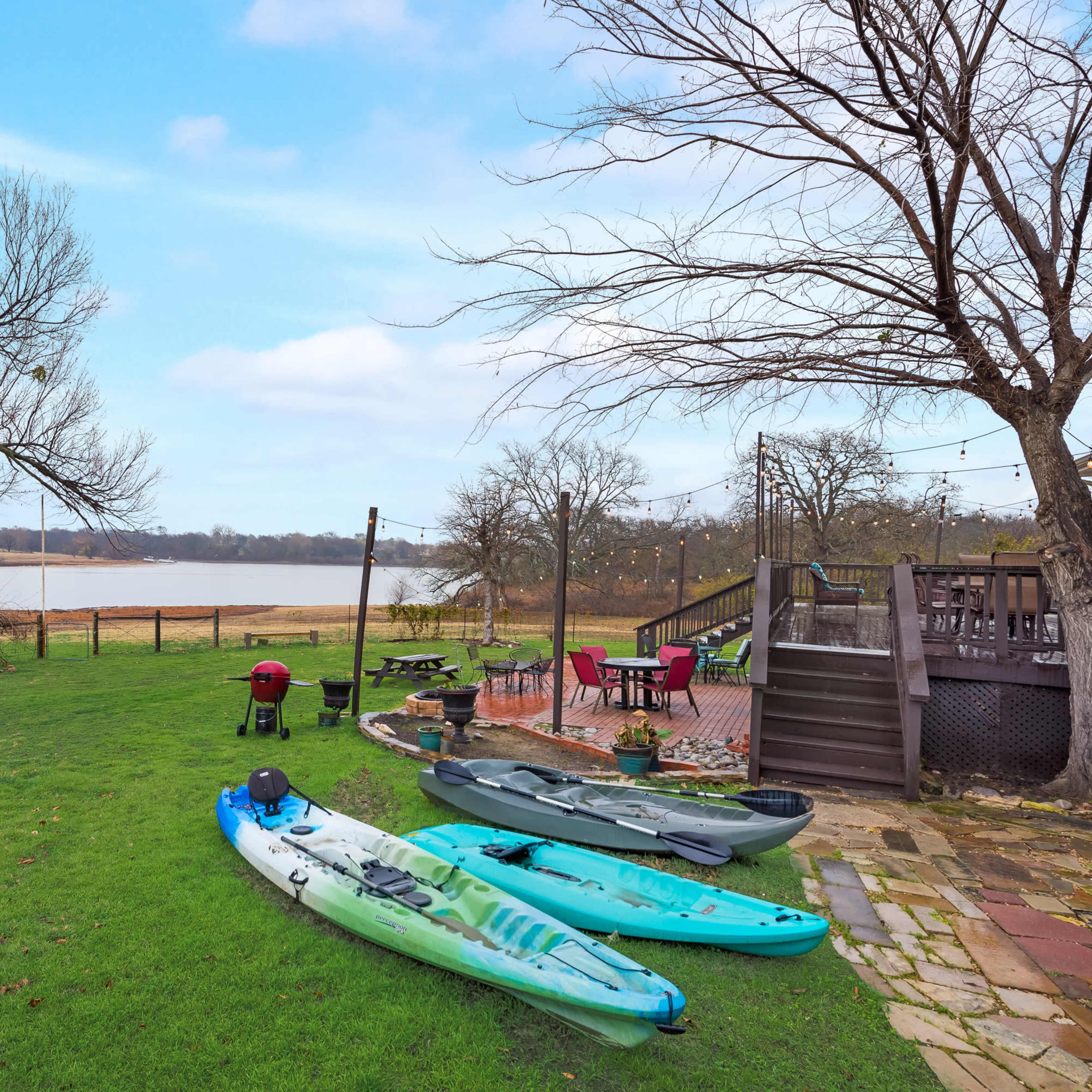 A grassy area by a river with kayaks on the shore, a picnic table, and a deck in the background.