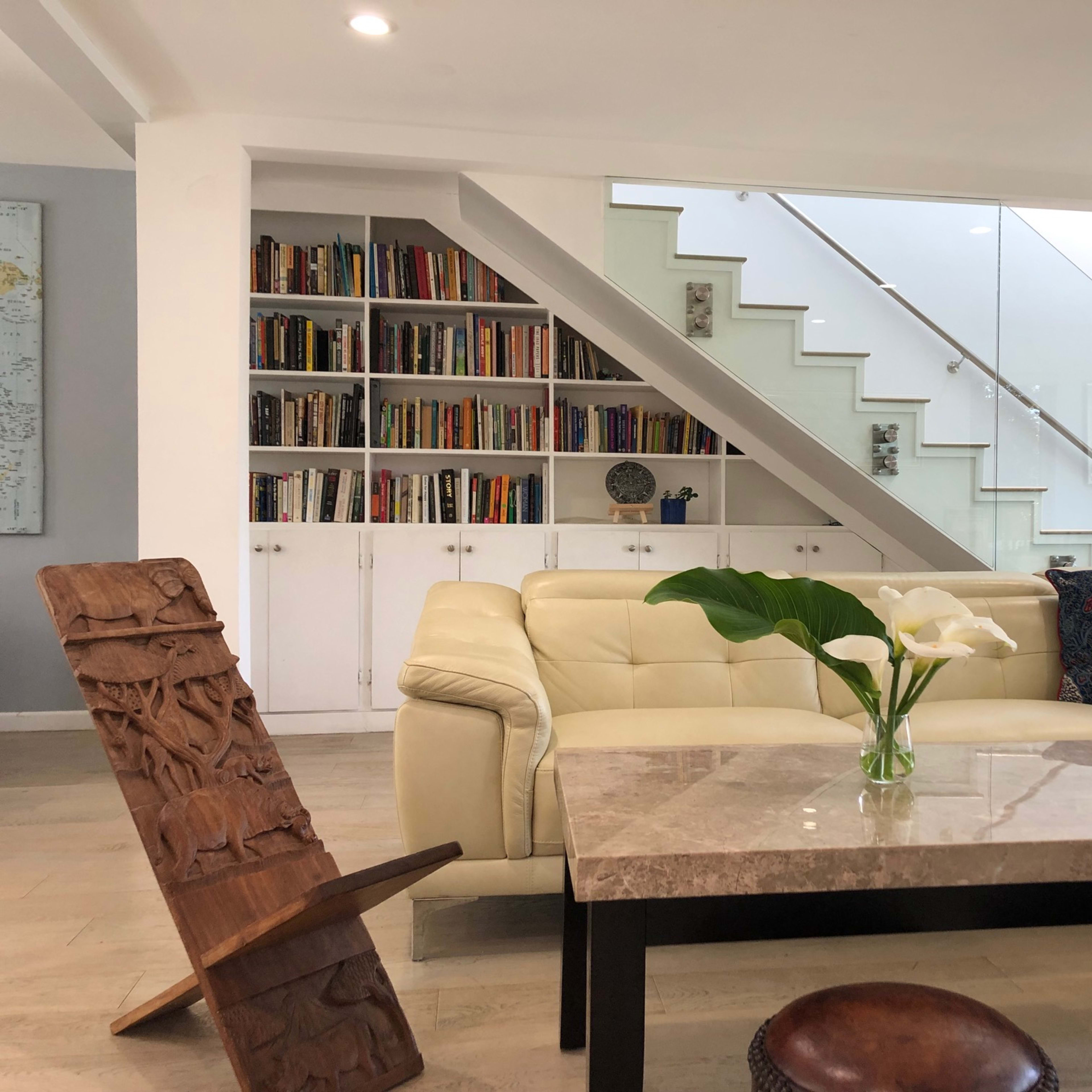 A modern living room with a cream-colored sofa, a marble coffee table, and a staircase leading to an upper level, alongside a bookshelf filled with books.