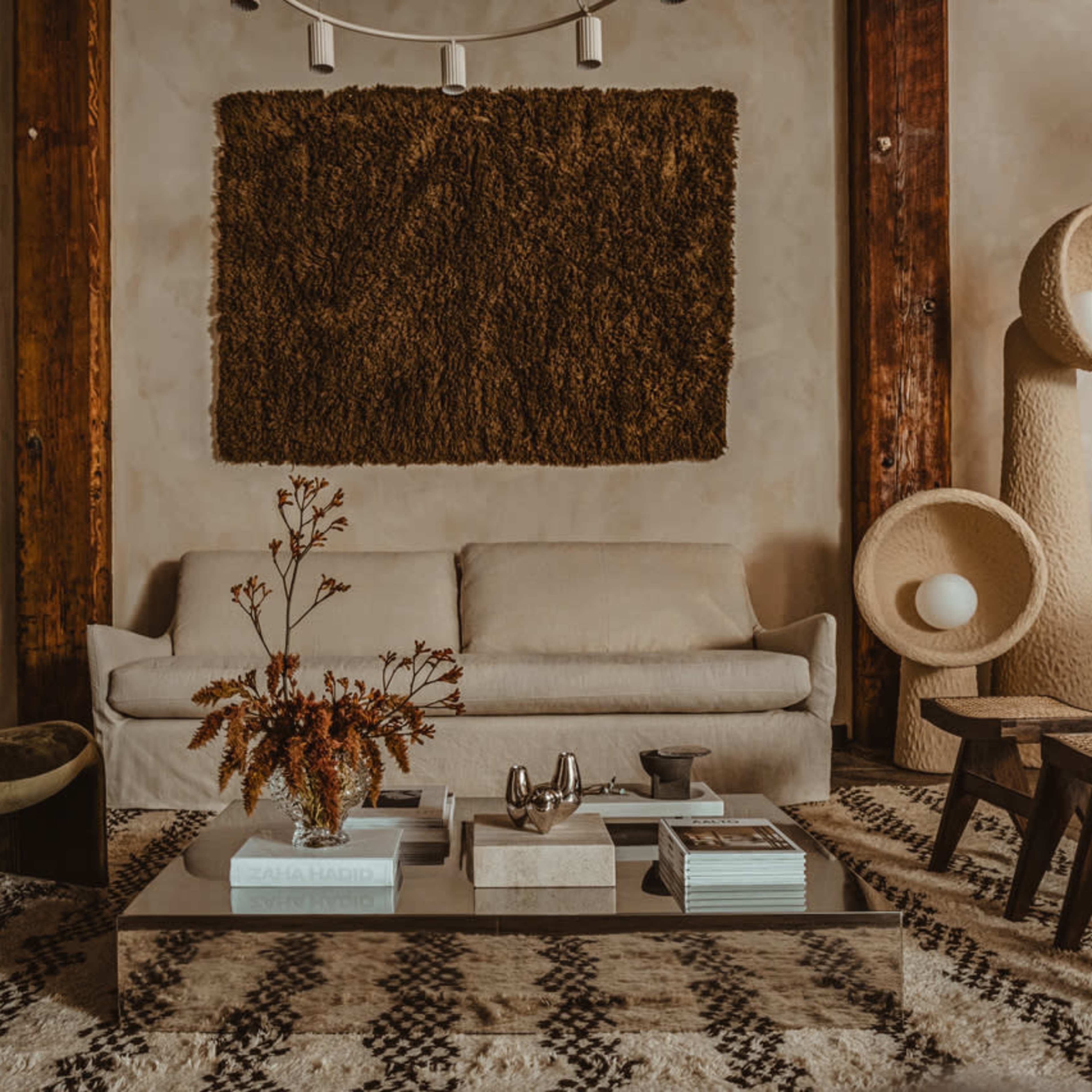A modern living room features a light-colored sofa, a glass coffee table with books and decor, and textured wall art, all set against warm, neutral tones.