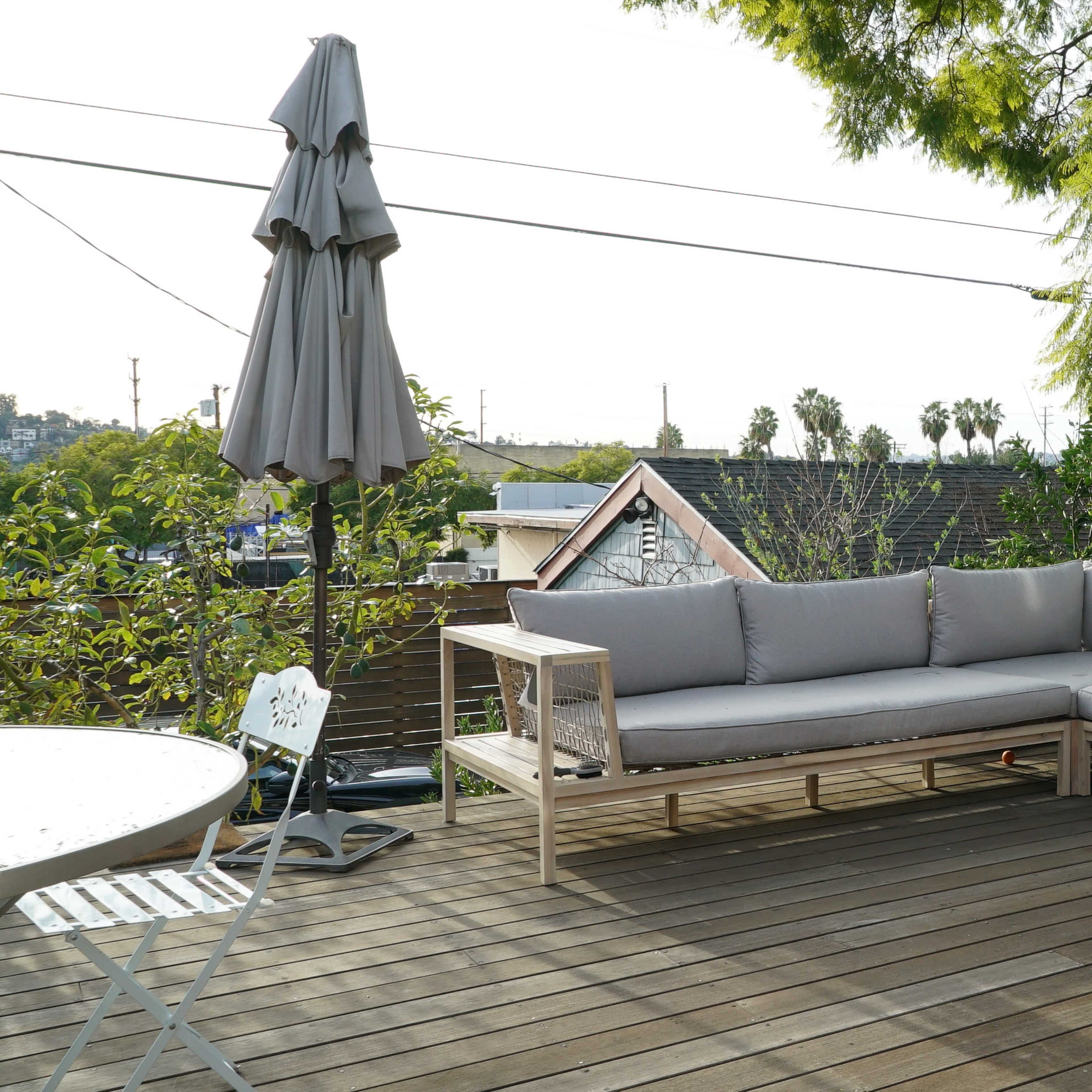 A light-colored sectional sofa with cushions is placed on a wooden deck surrounded by greenery and a sun umbrella.