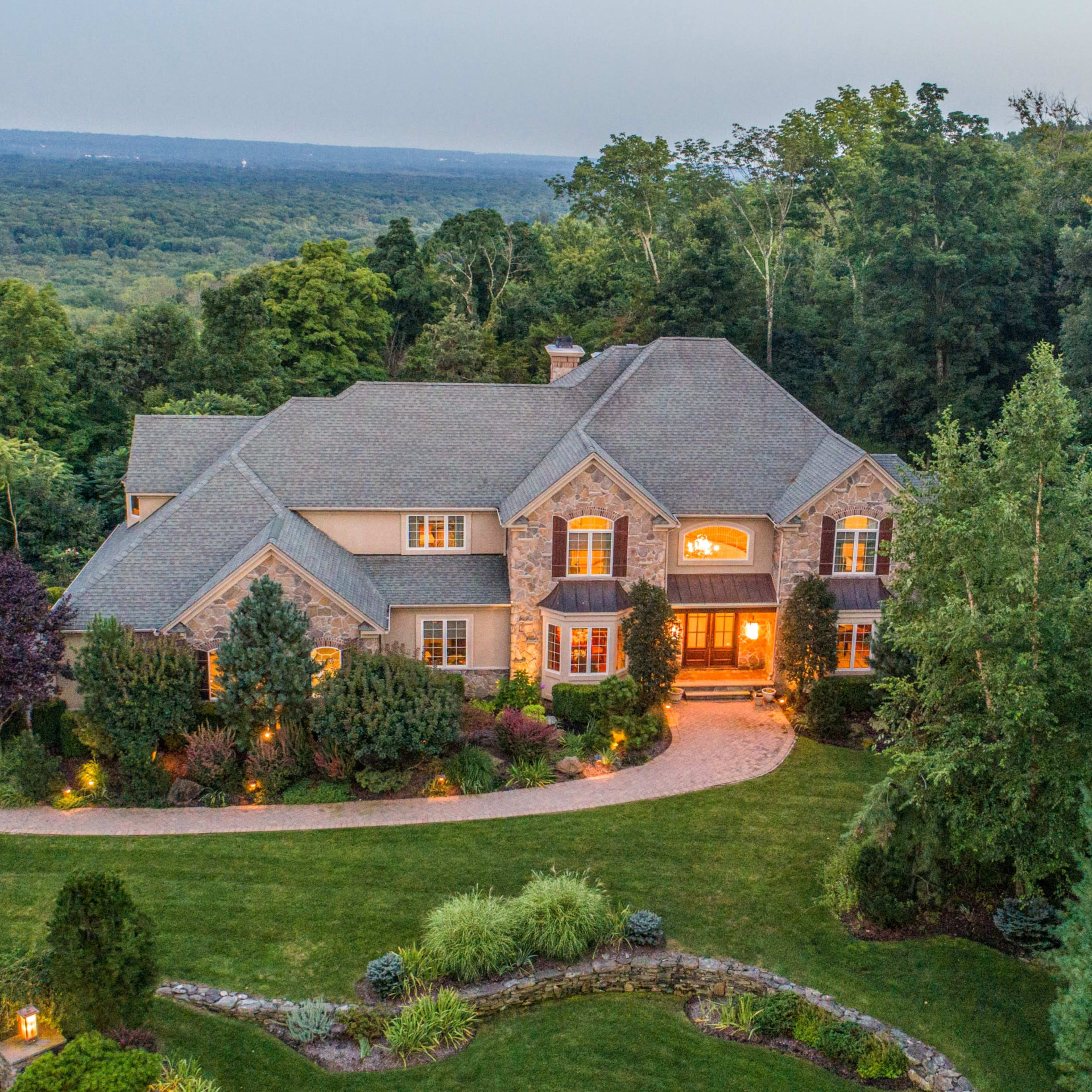 A large, multi-story stone and shingle house is set on a landscaped property surrounded by trees and hills, with warm lights illuminating the entrance.