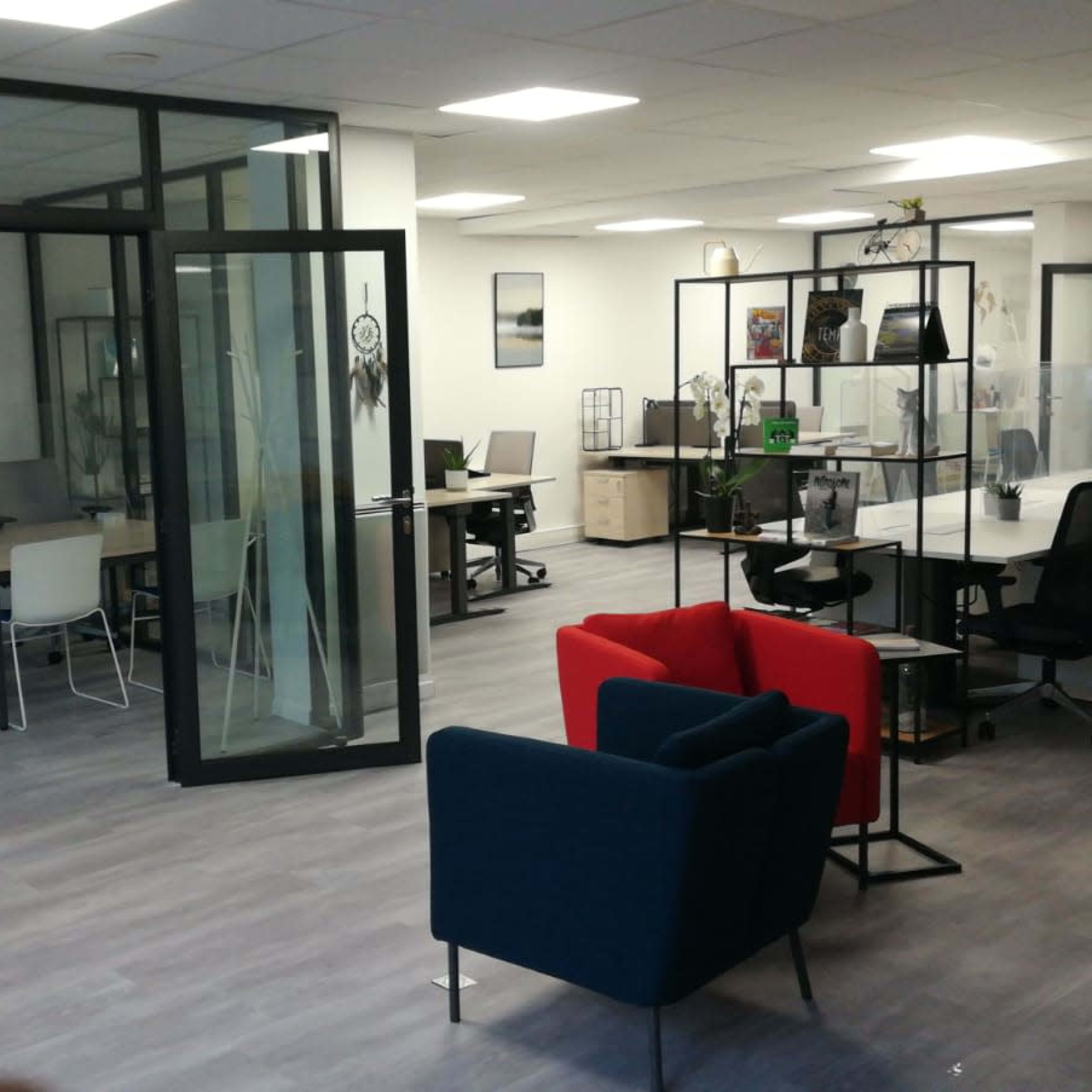 A modern office space featuring glass partitioned areas, several desks with chairs, and a seating area with red and blue couches.