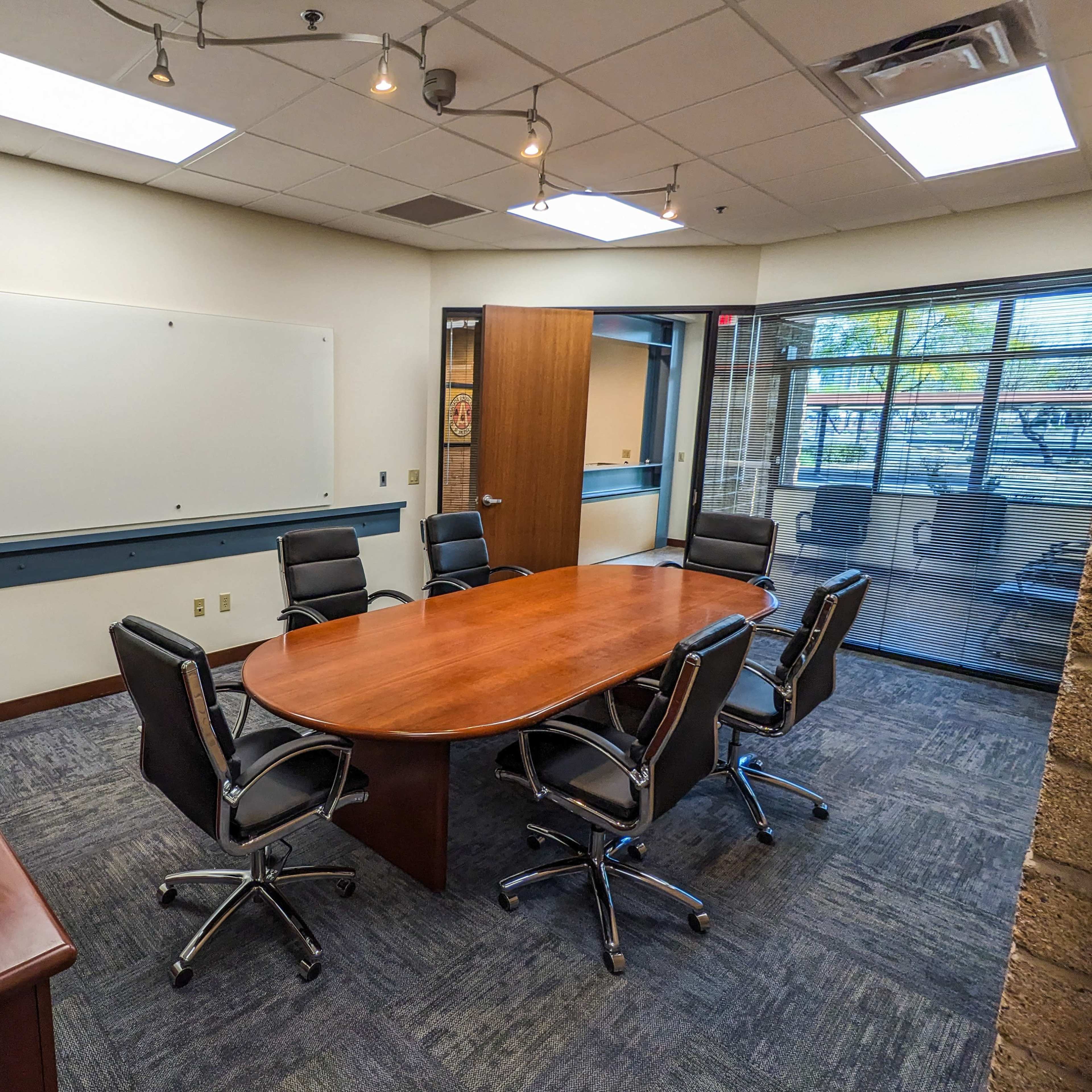 A conference room features a large wooden table surrounded by six black chairs, with a whiteboard and a TV screen on the wall.