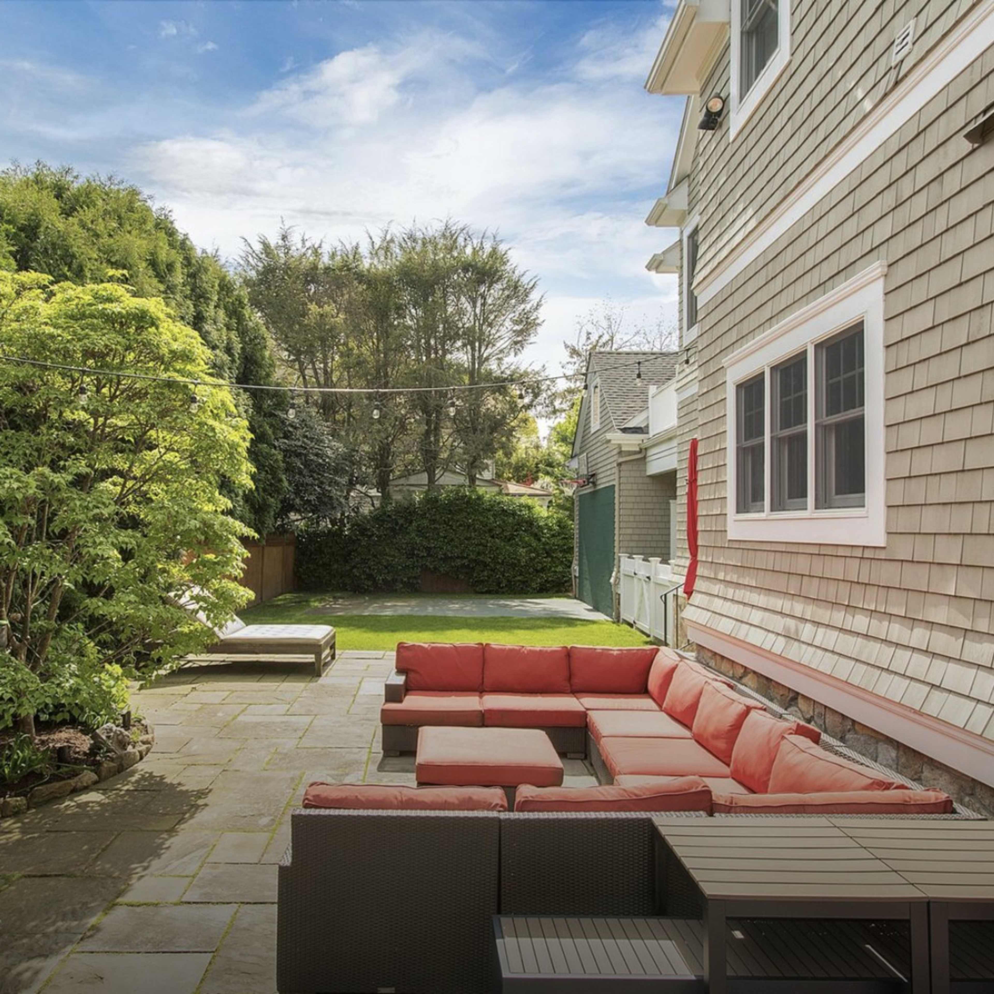 A patio features a large sectional sofa with red cushions, surrounded by greenery and stone pathways.