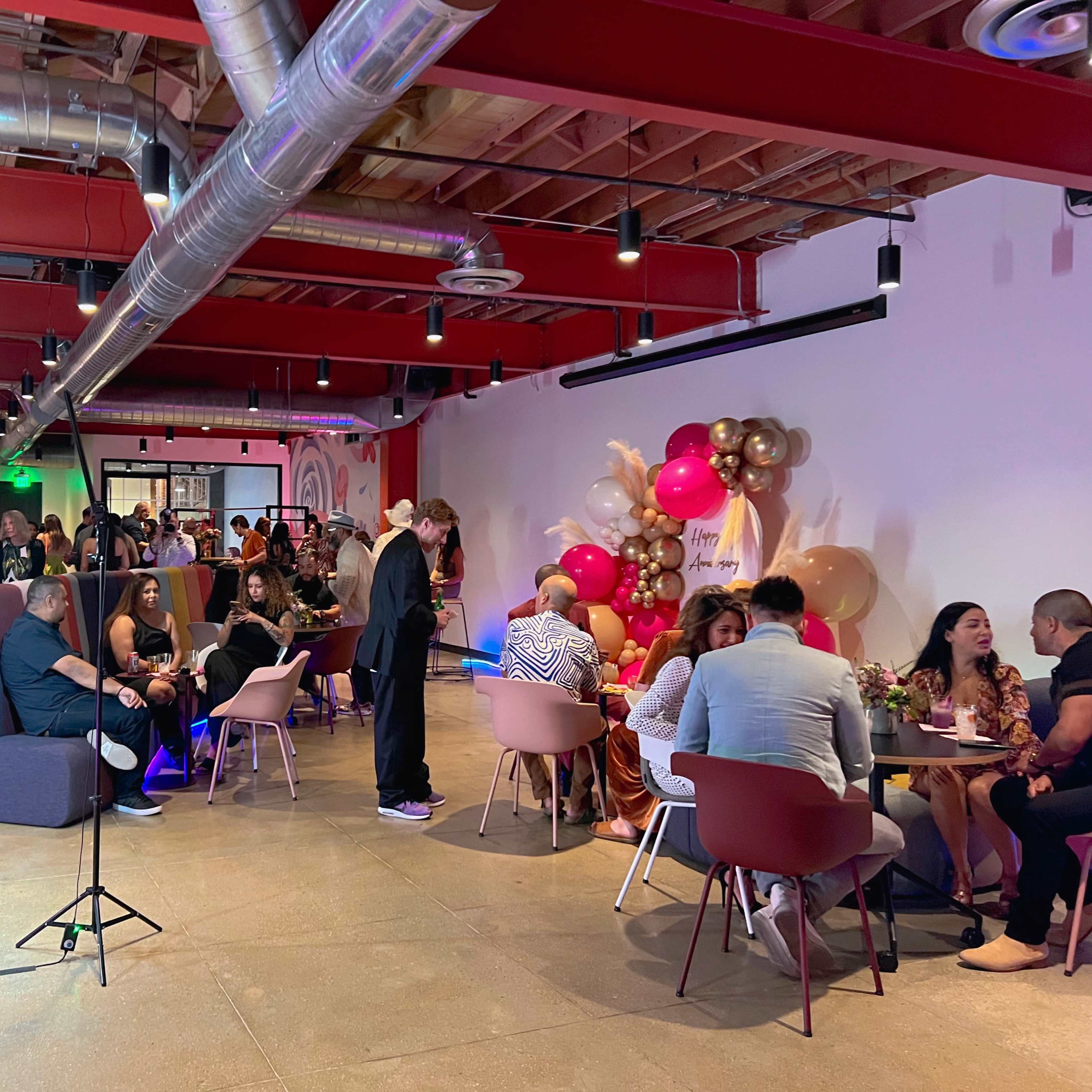 A crowded event space features guests seated at various tables, mingling and enjoying food and drinks amid colorful decorations.