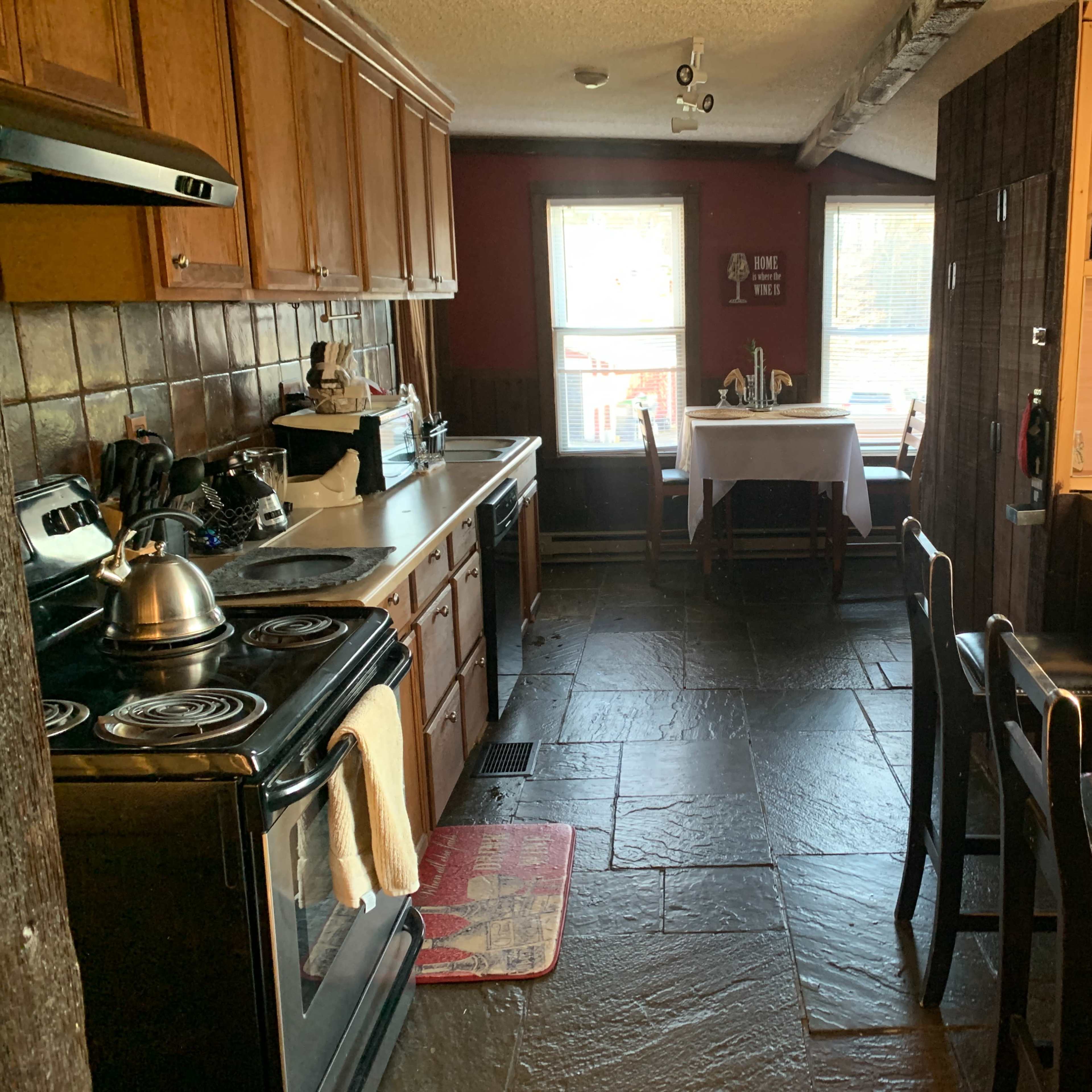 A kitchen features wooden cabinets, a stove, and a stone floor, with a table and chairs visible in the adjoining dining area.
