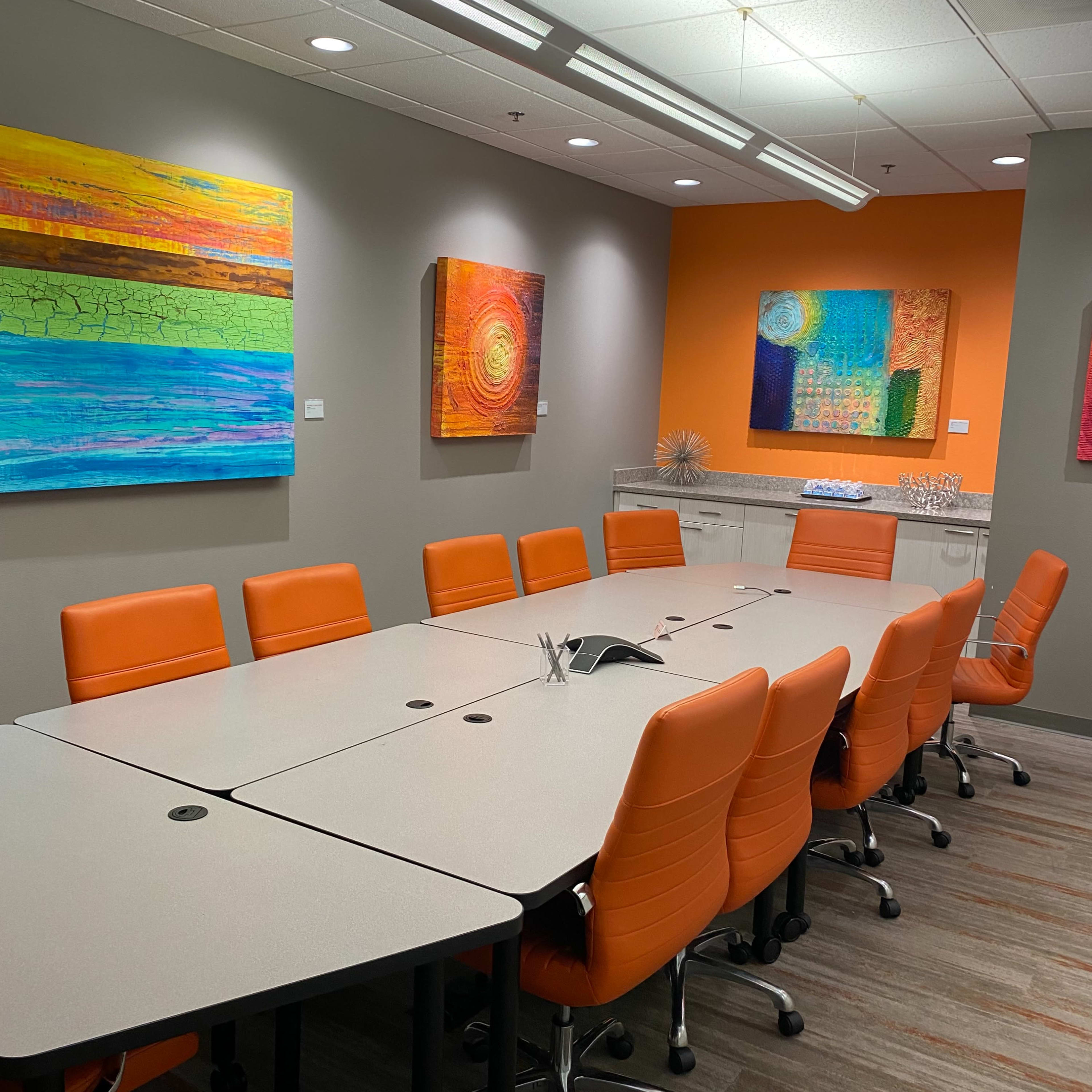 A modern conference room features a long table surrounded by orange chairs, with colorful abstract art displayed on the walls.