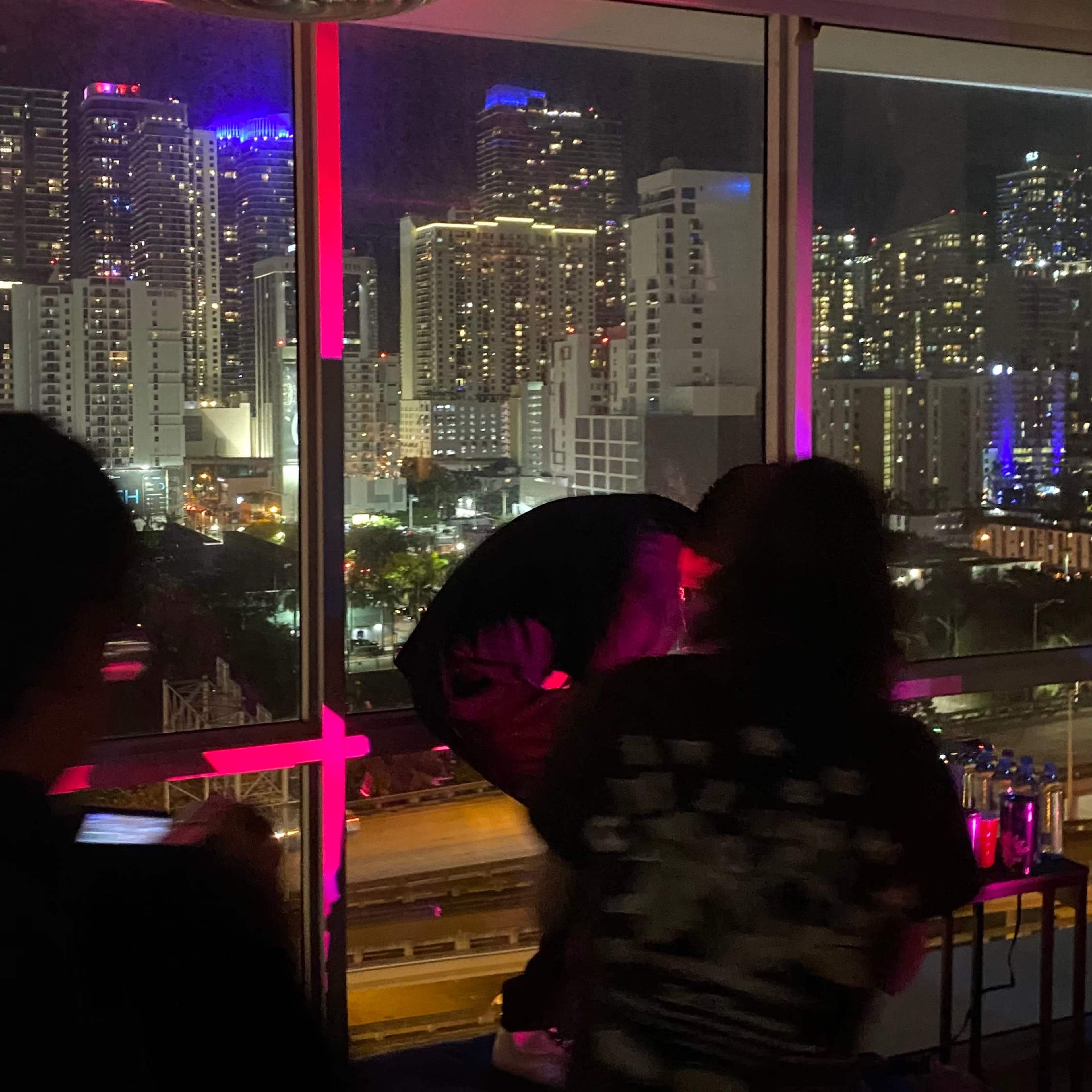A group of people is gathered in a brightly lit room with city skyline views, featuring high-rise buildings illuminated at night.