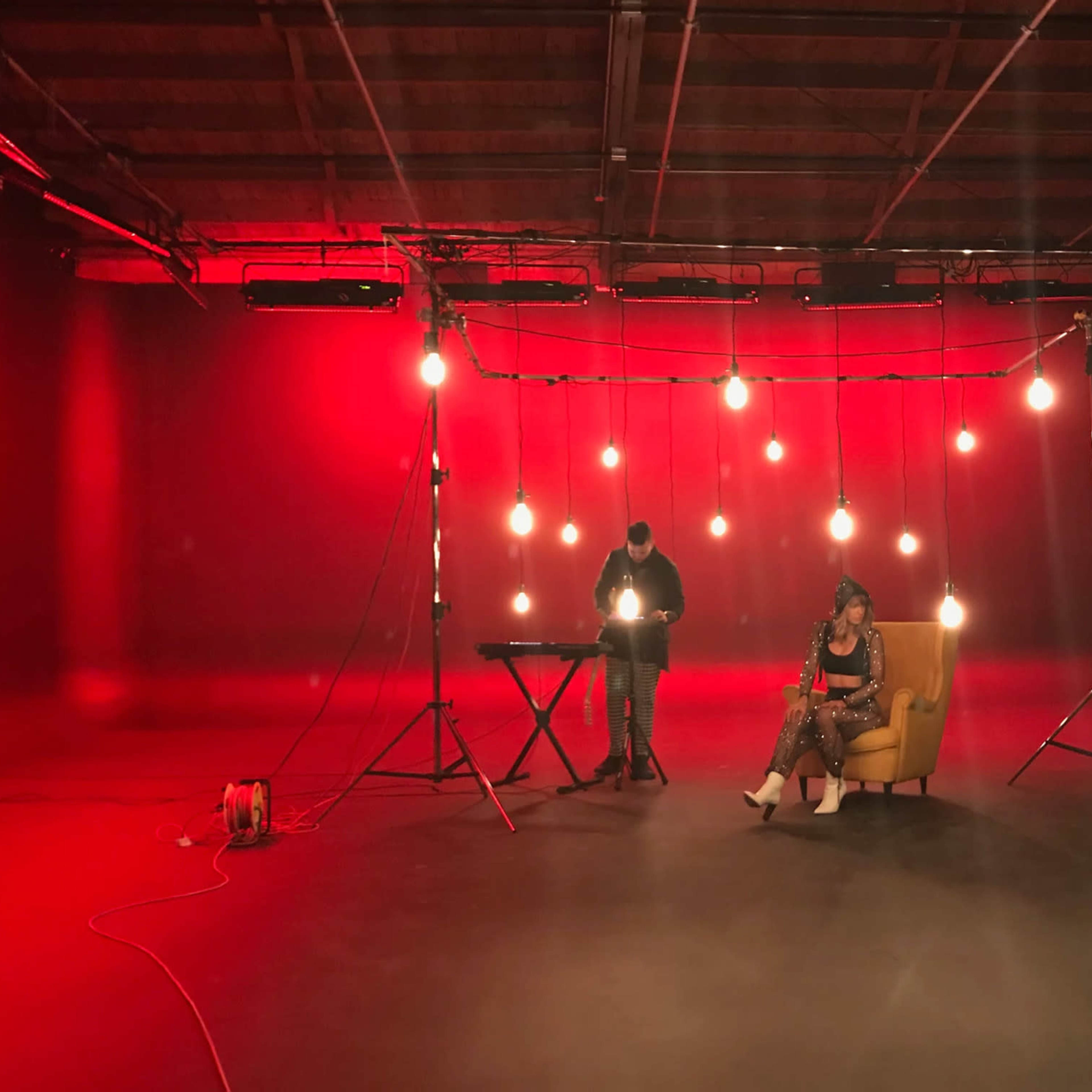 A woman sits in a yellow chair under hanging lights while a man stands at a keyboard in a red-lit studio.
