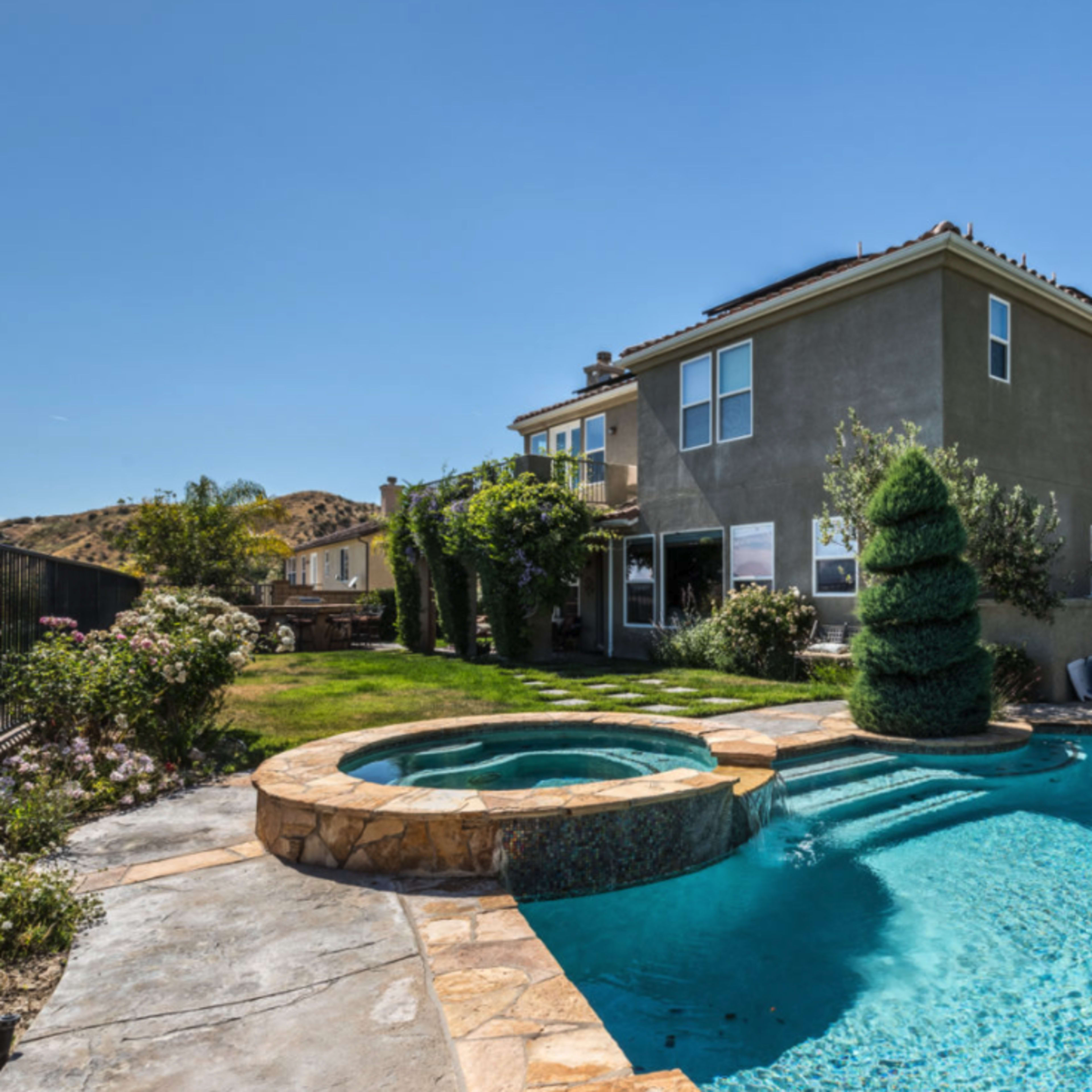 A two-story house with a fenced backyard features a swimming pool and a spa surrounded by landscaping and hills in the background.