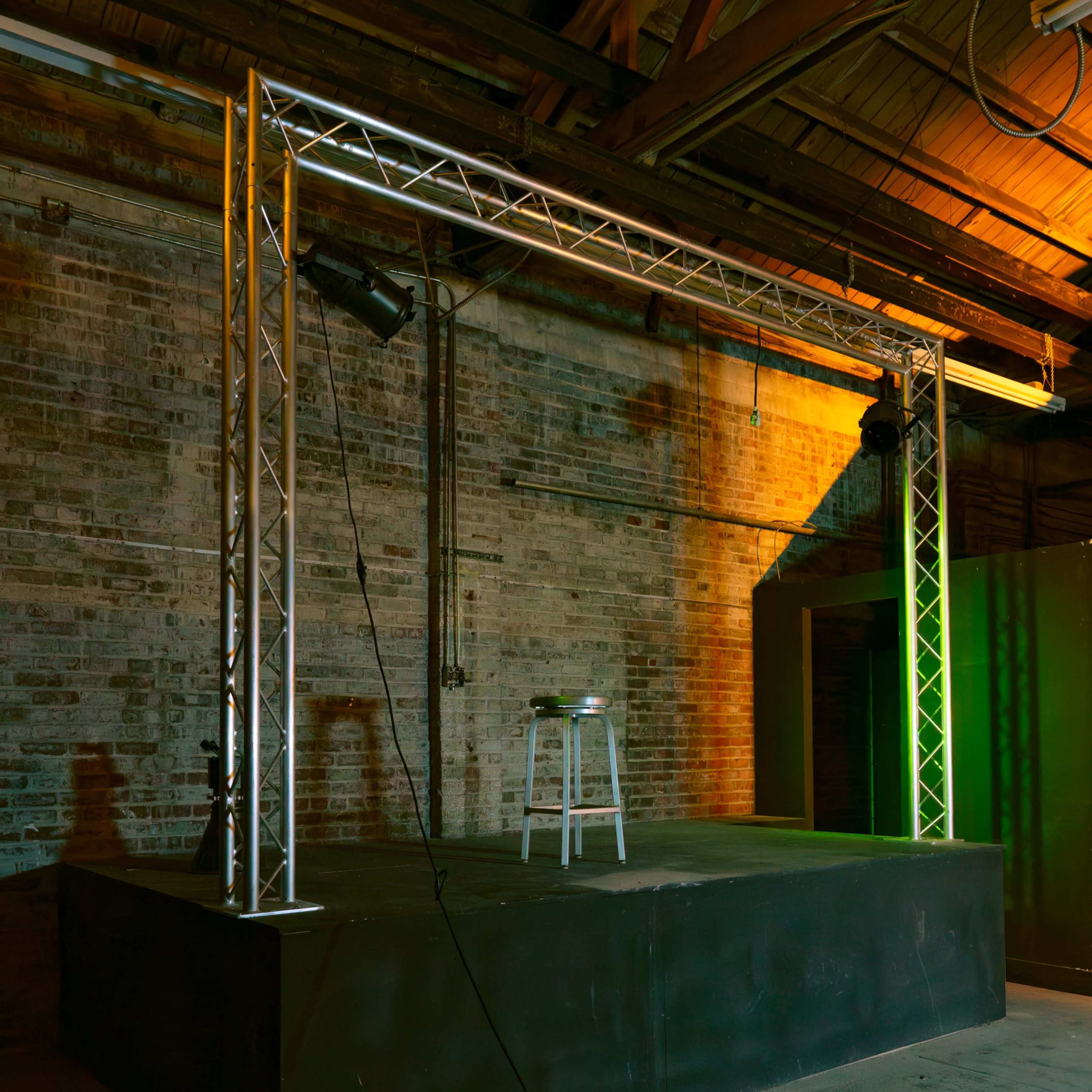 A raised stage with a stool is set up in a spacious industrial room with exposed brick walls and overhead lighting.