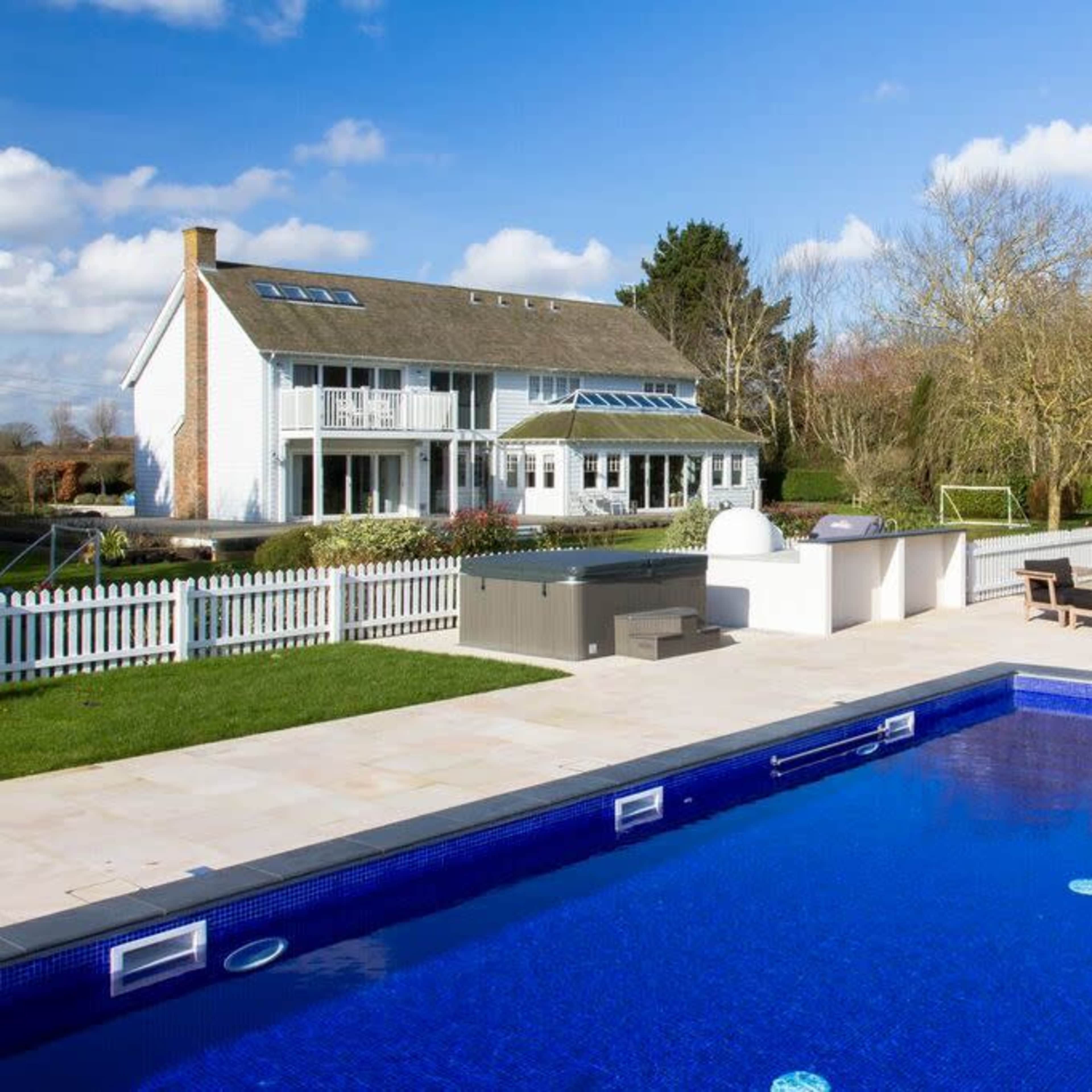 A backyard with a blue swimming pool, a large house in the background, and a patio area with seating and an outdoor kitchen.