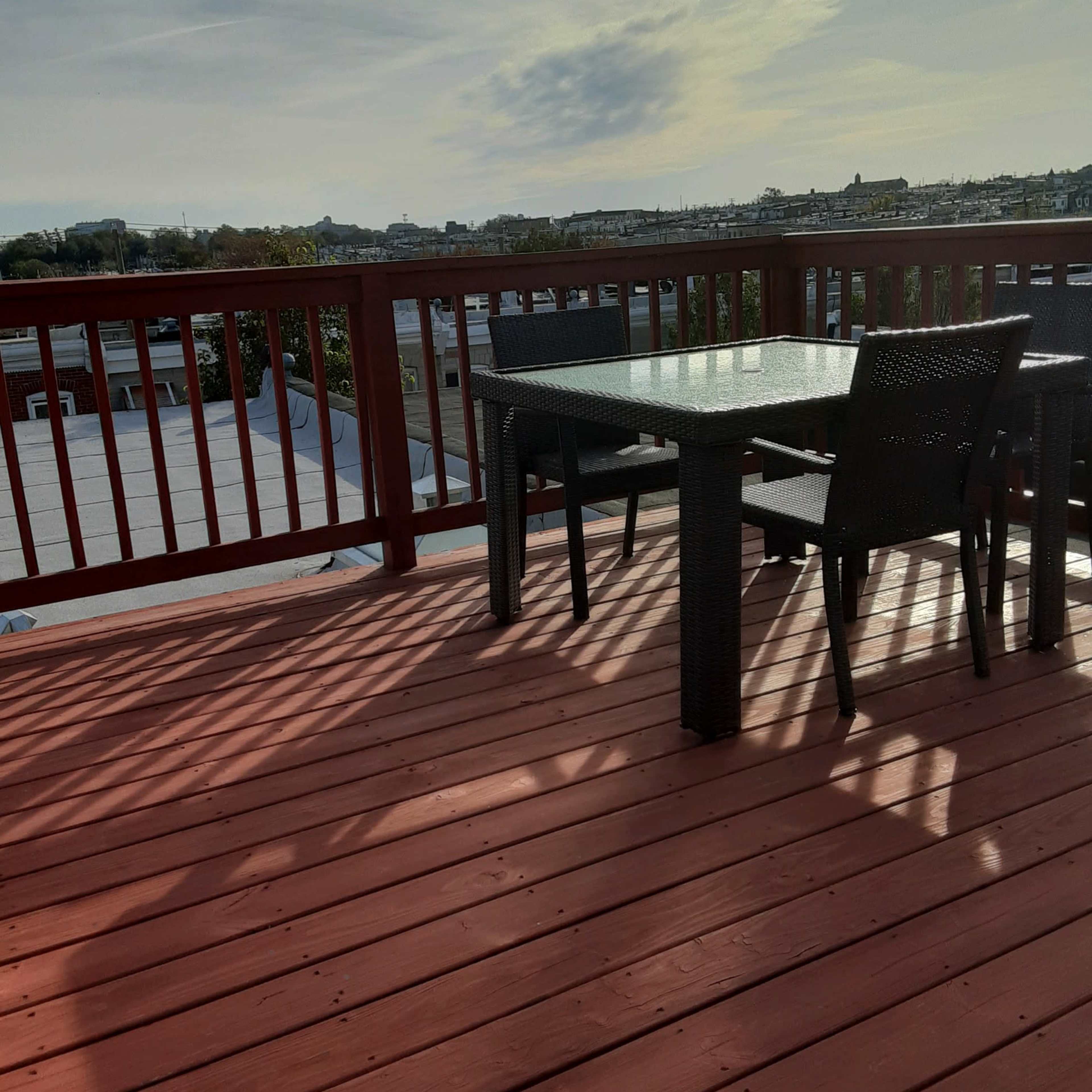 A wooden deck features a glass-top table and chairs, overlooking a distant landscape under a partly cloudy sky.