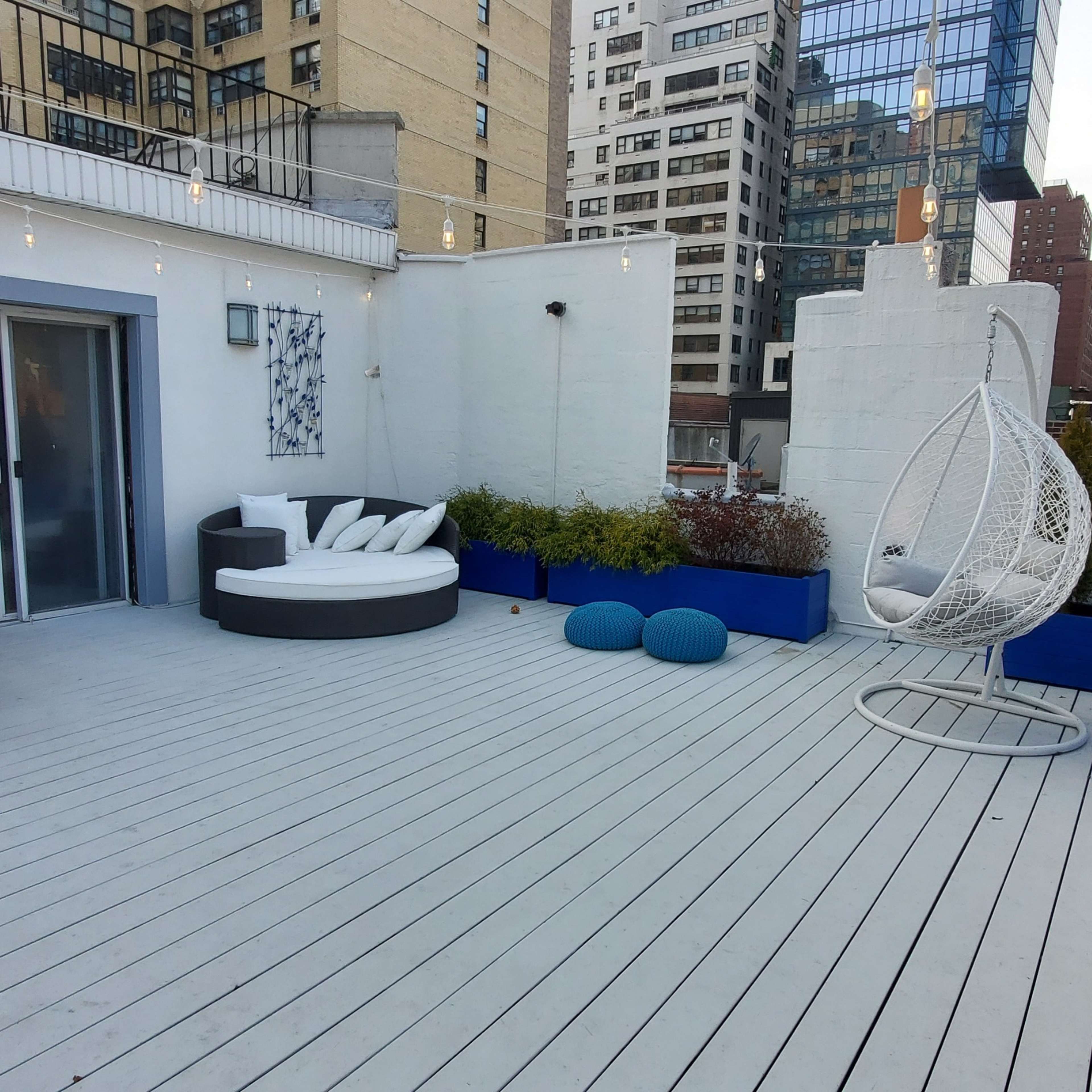 A spacious rooftop terrace featuring seating areas, colorful planters, and city buildings in the background.