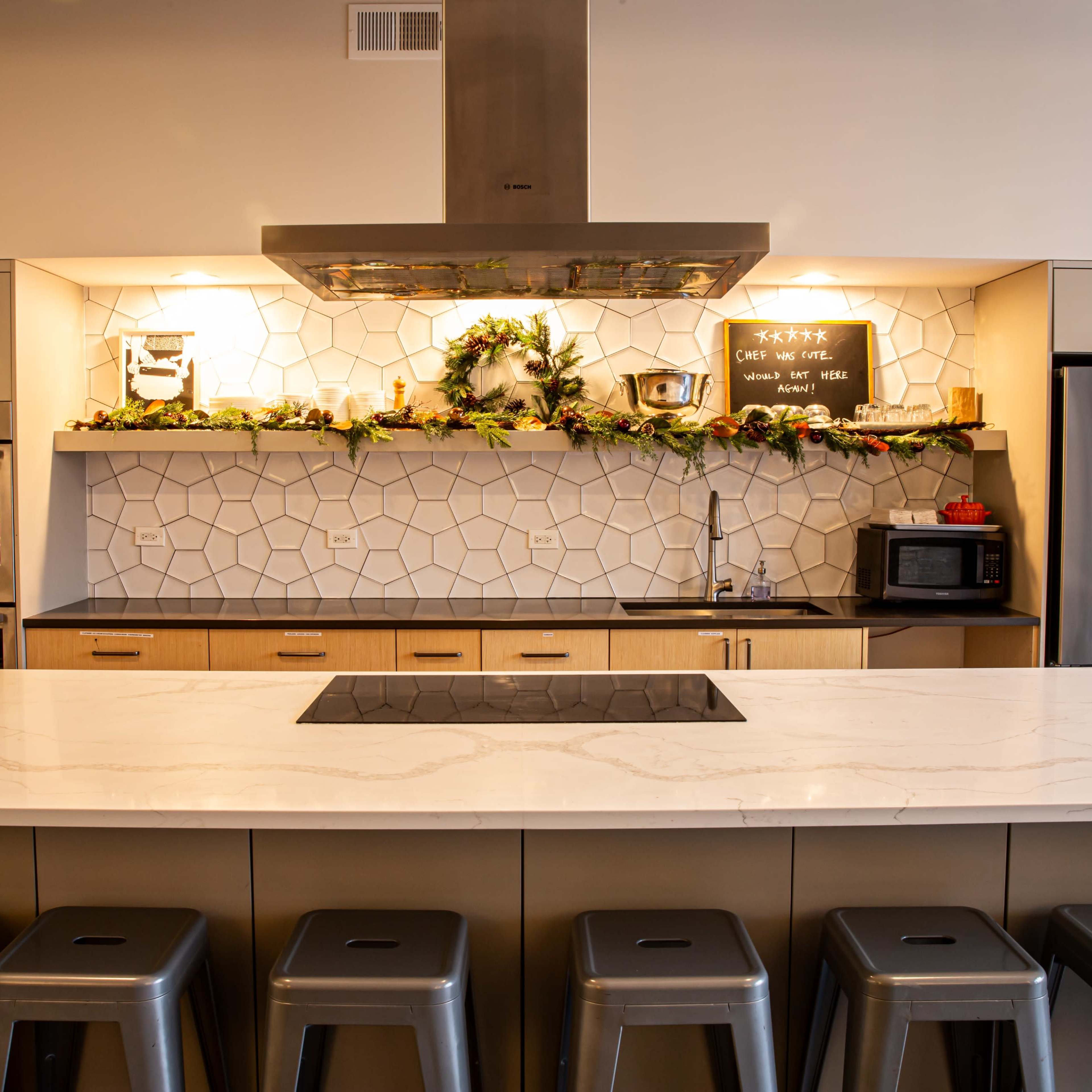A modern kitchen with a large island, featuring a geometric tiled backsplash, stainless steel appliances, and metal barstools lined up along the counter.