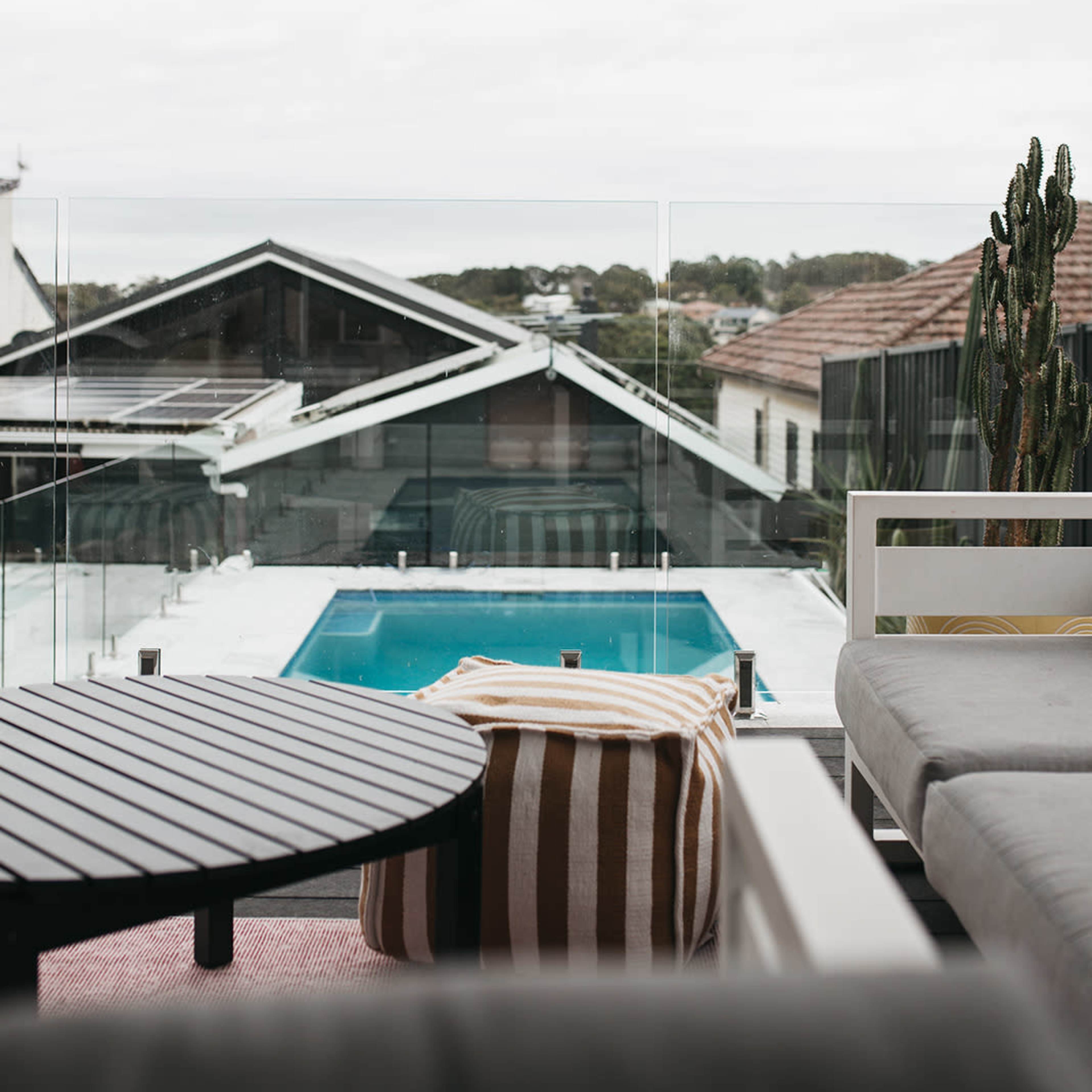 A modern outdoor patio featuring a table and seating area, overlooking a swimming pool surrounded by buildings and greenery.