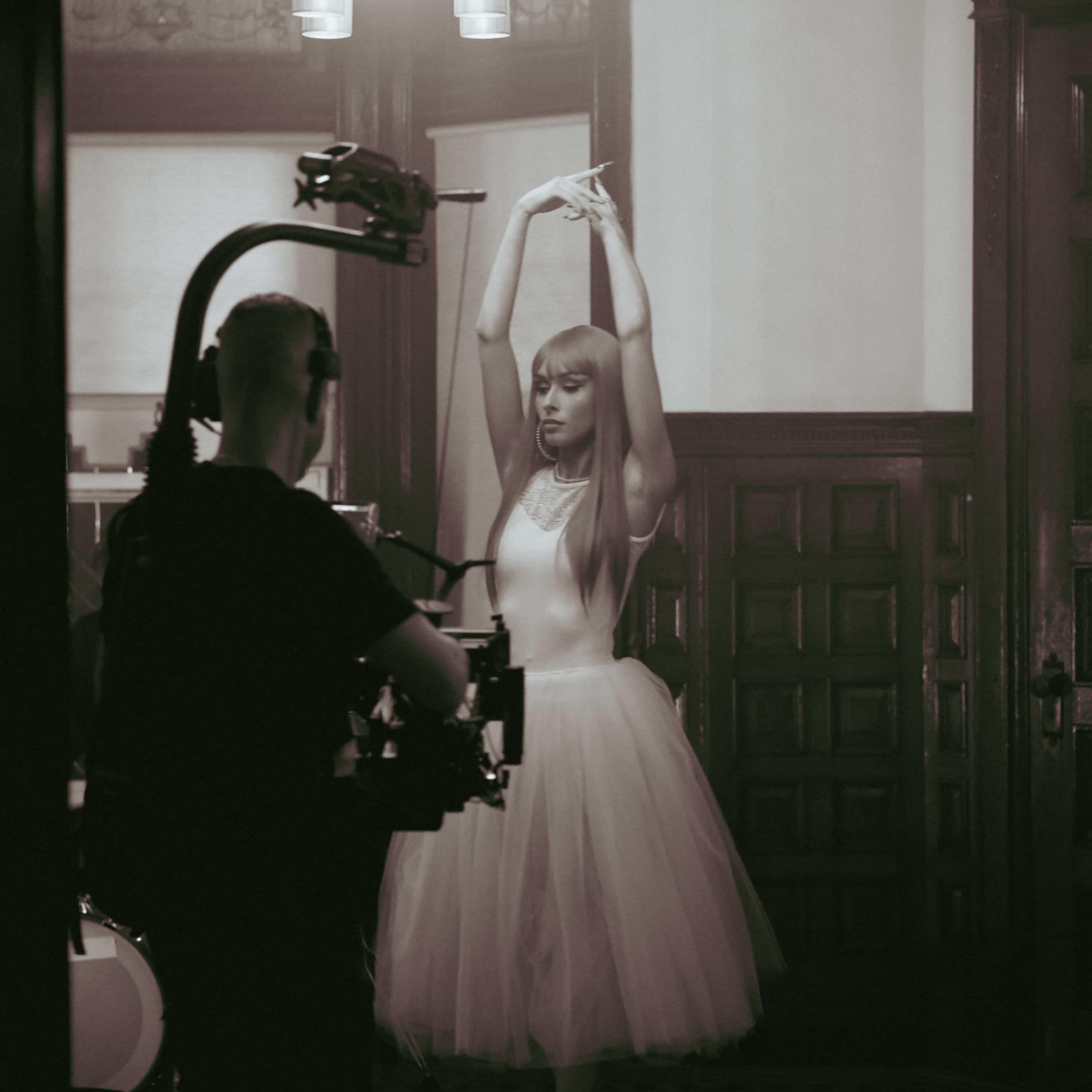 A dancer in a tutu strikes a pose while being filmed by a cameraman in a dimly lit room.