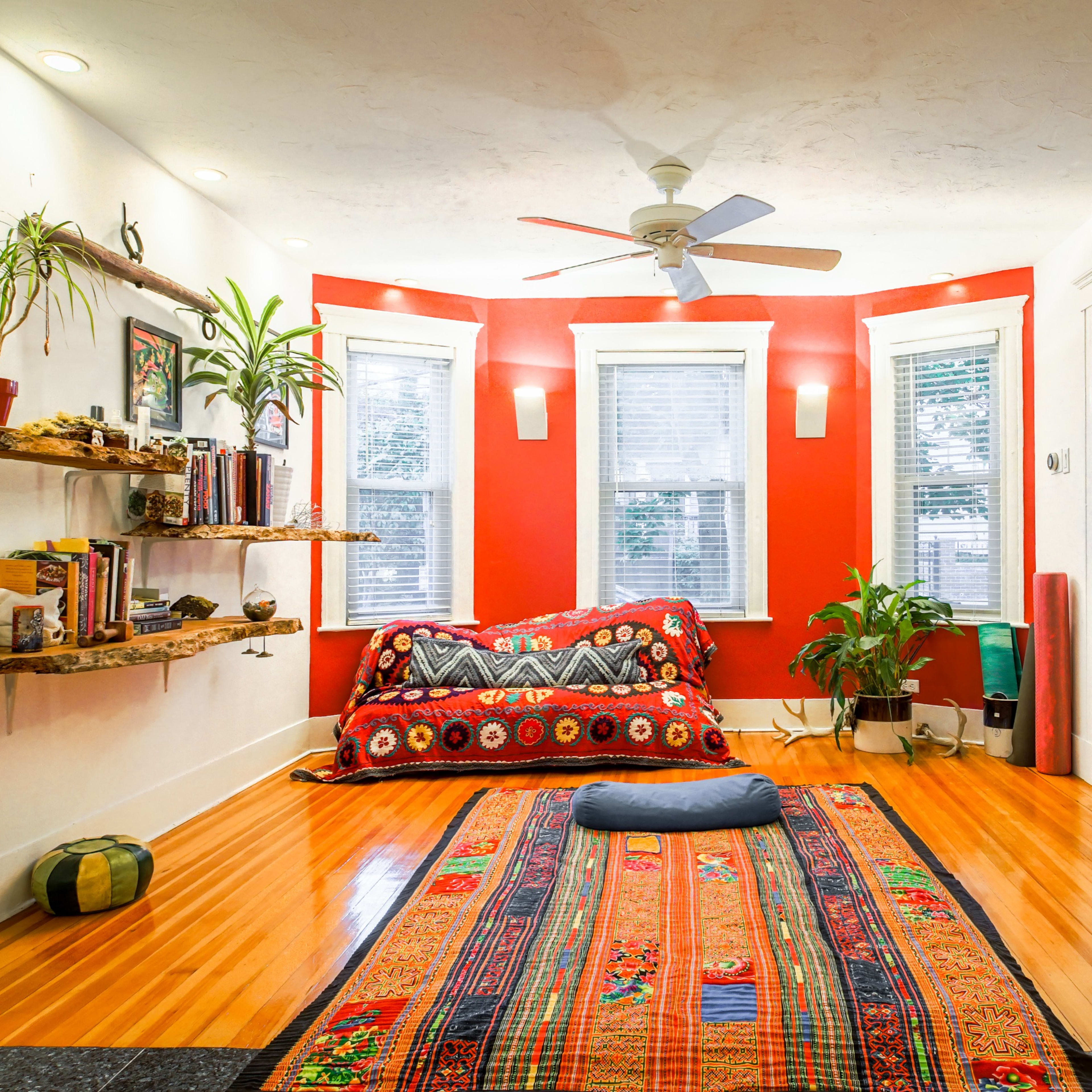 A brightly colored living room with a red accent wall, a wooden floor, a patterned rug, a shelf with decorative items, and a door leading to another room.