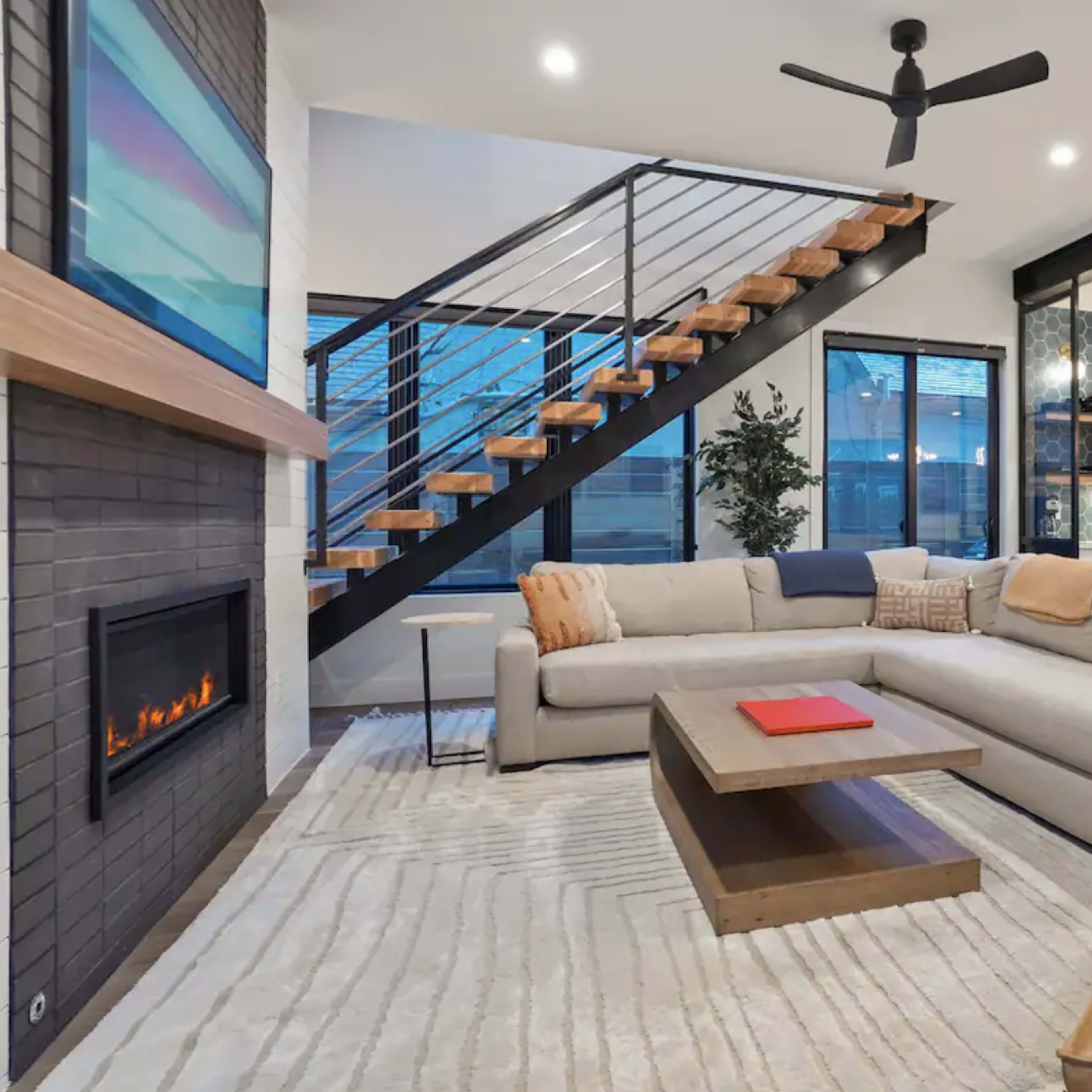 A modern living room with a large sectional sofa, a coffee table, a fireplace, and an open staircase leading to an upper level.