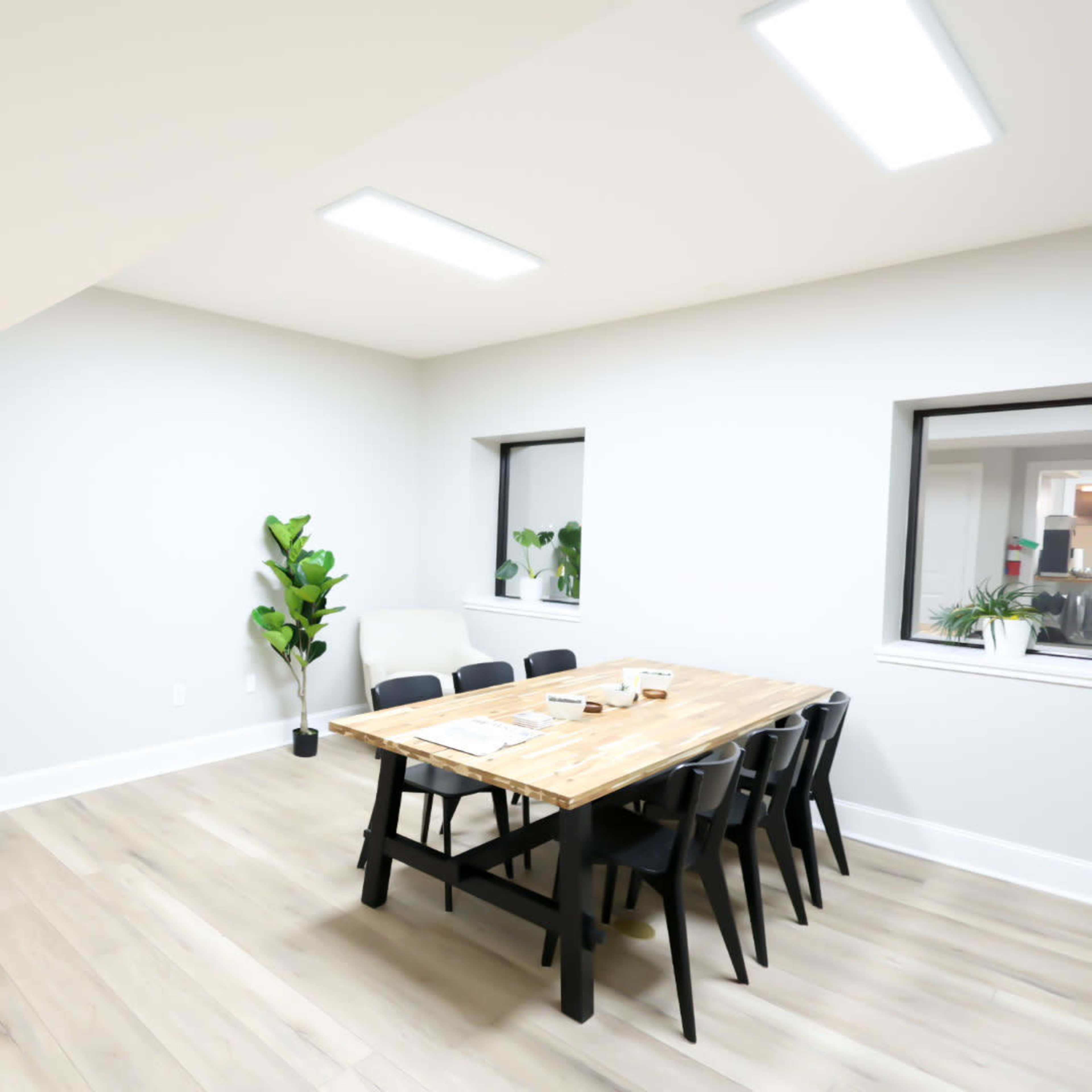 A modern hallway leading to a conference room with a wooden table and black chairs, accompanied by a potted plant by the window.
