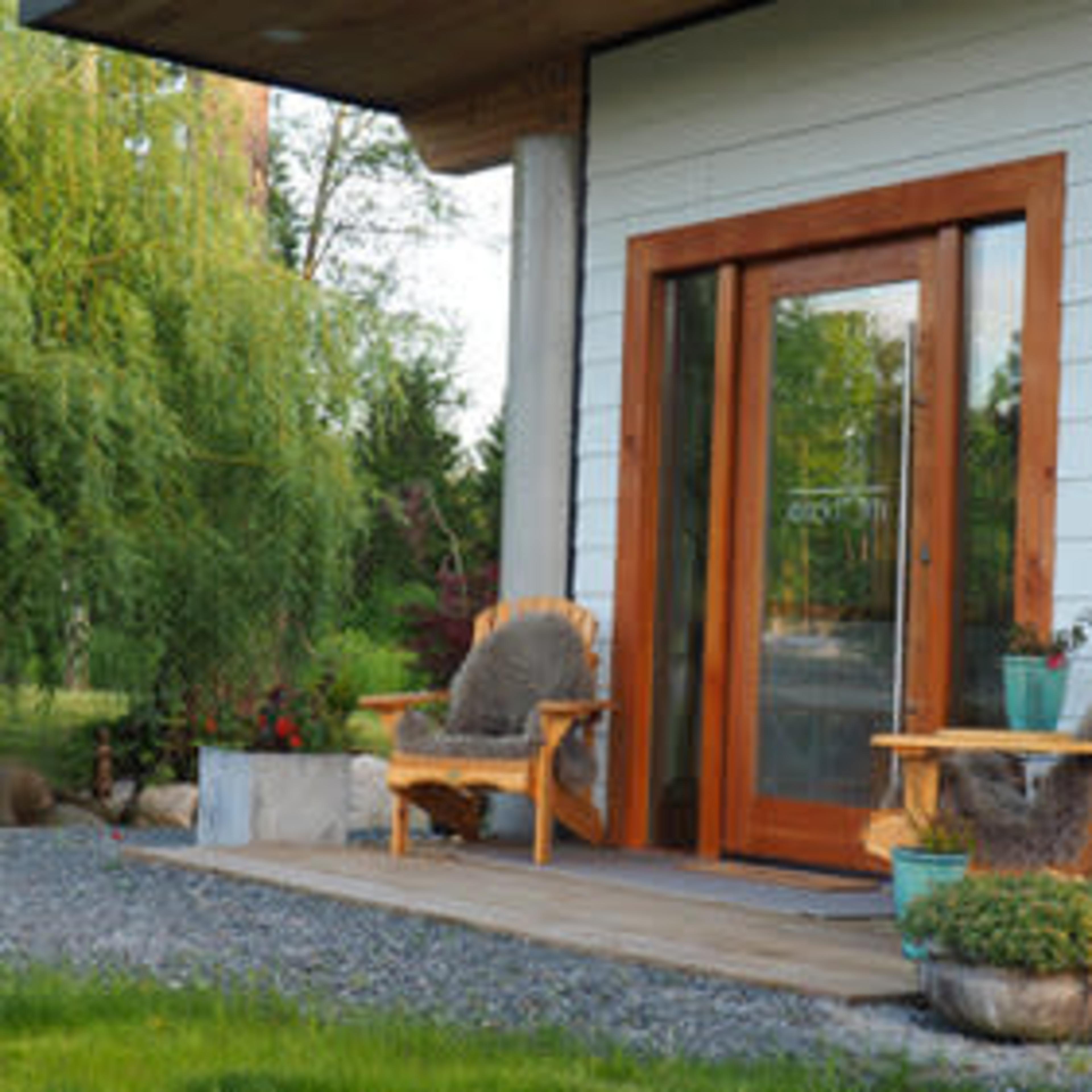 A wooden porch with two chairs and a potted plant sits beside a grassy area and trees.