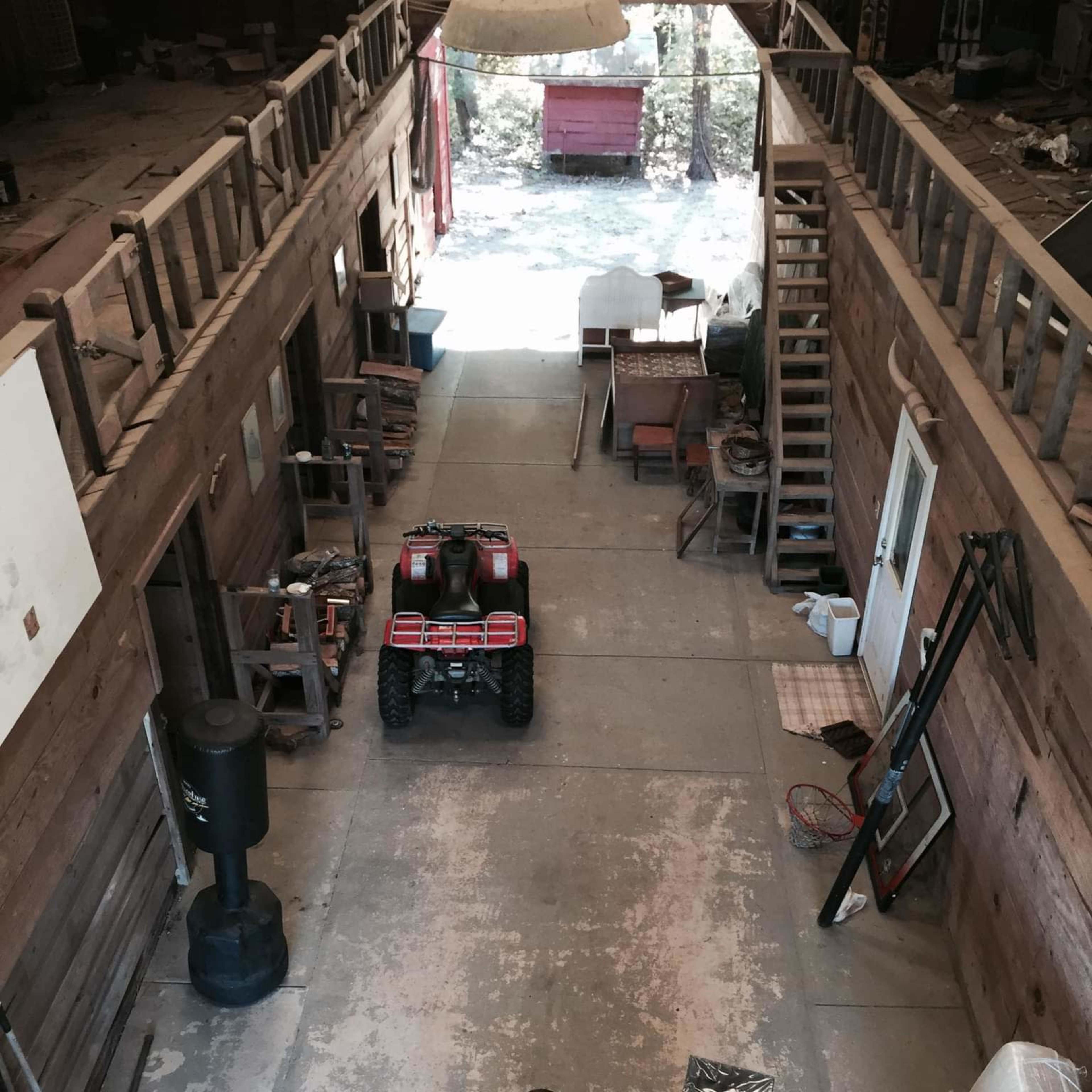A spacious, unfinished interior of a barn-like structure with an ATV parked in the center, surrounded by various items and wooden beams overhead.