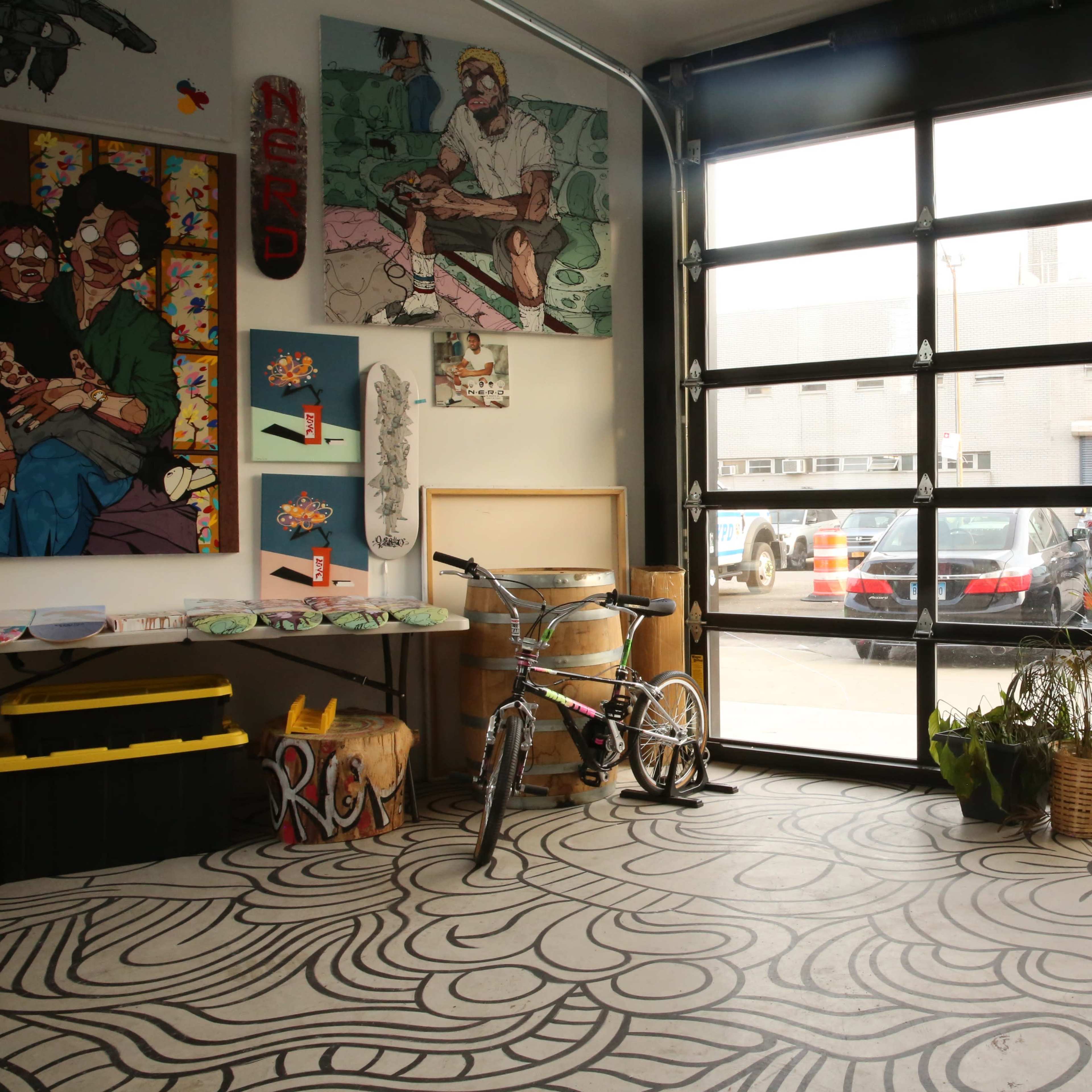 An artist's studio with a large garage door, artwork on the walls, a bicycle, a blank easel, and a patterned floor.