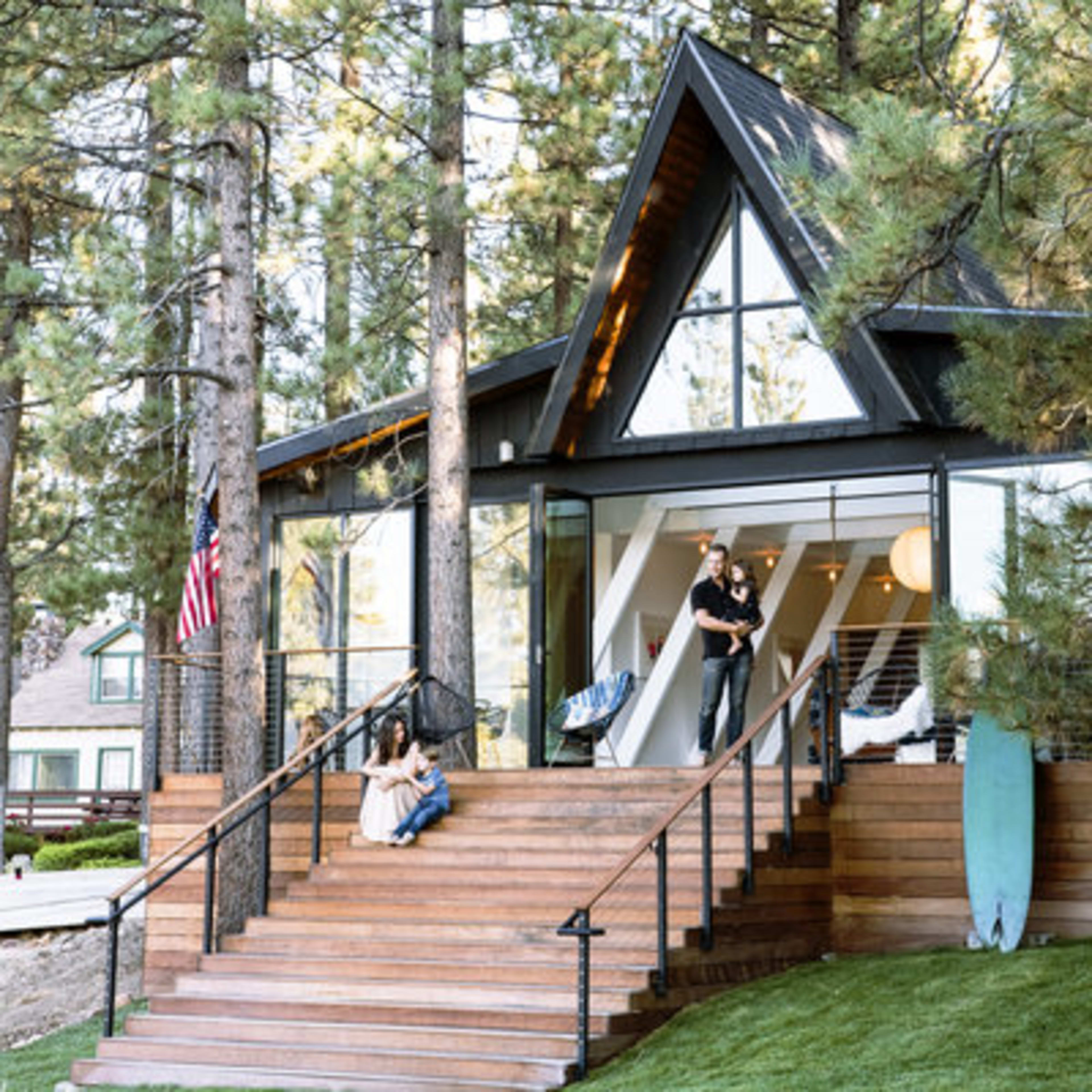 A modern A-frame house with large windows is surrounded by tall trees, featuring a wooden staircase leading to a porch where two people are engaged in conversation.