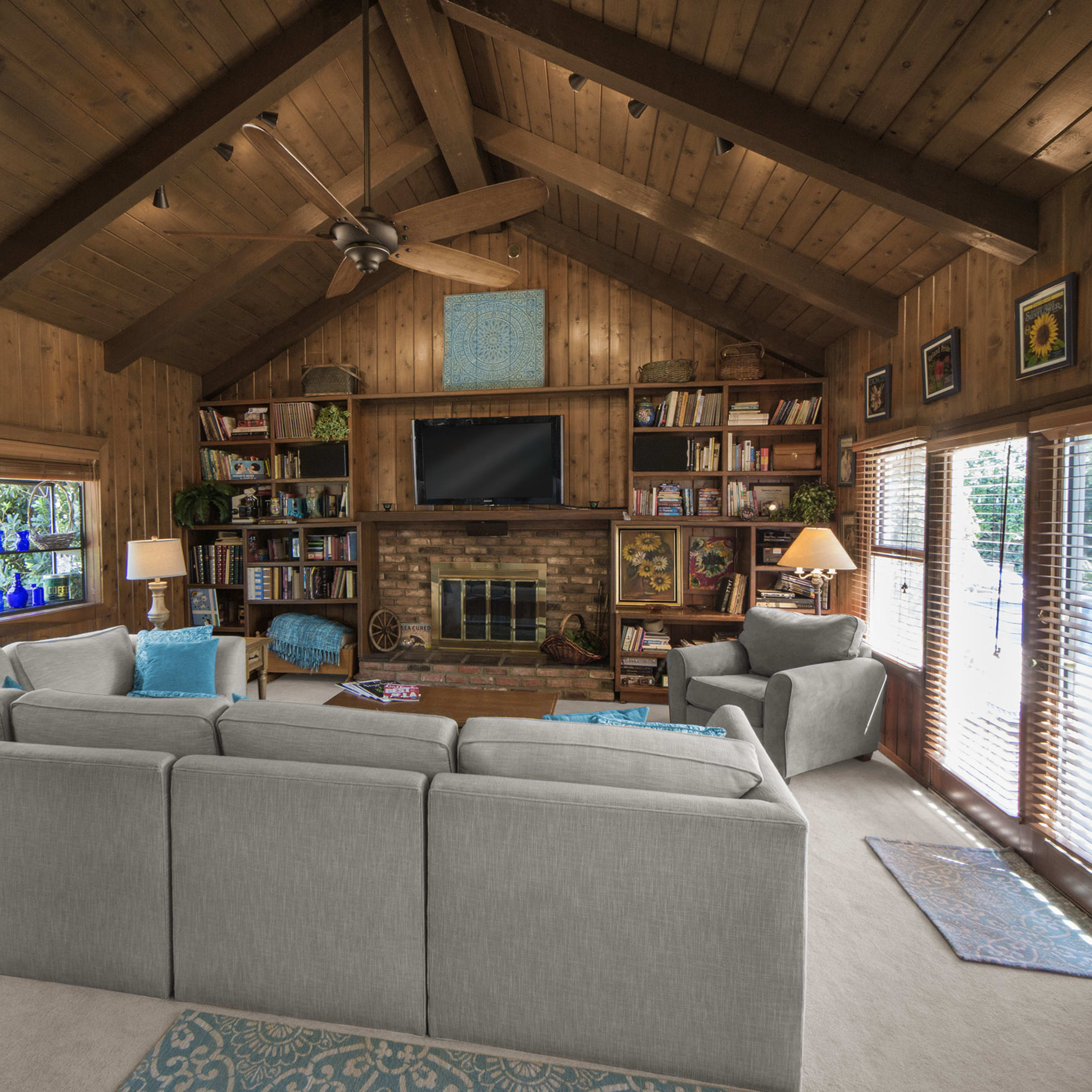 A rustic living room with wooden walls and a large couch facing a brick fireplace and a television, flanked by bookshelves and large windows.